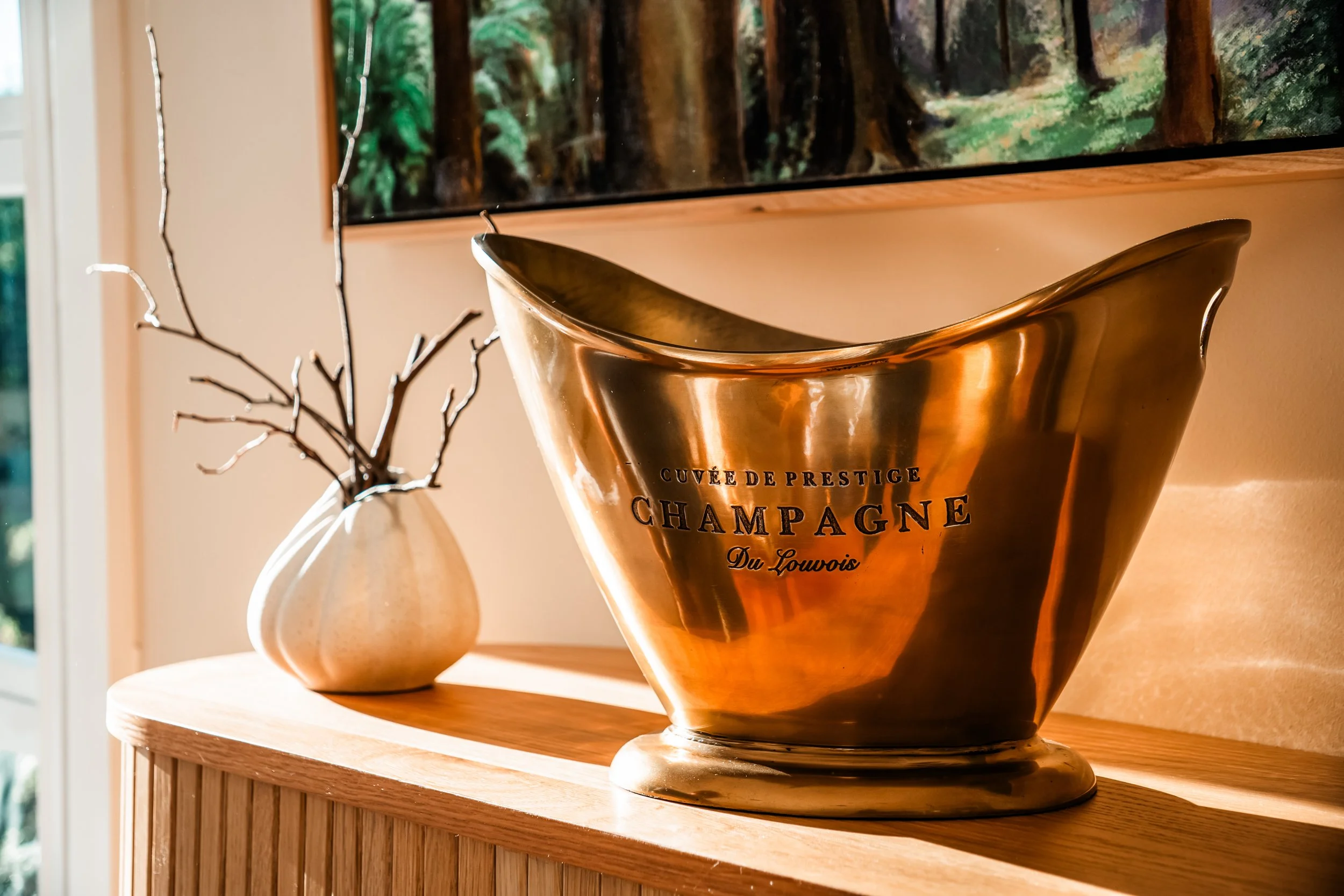 A large shiny gold champagne bucket with the words "CUVÉE DE PRESTIGE CHAMPAGNE Du Louwis" printed on it, placed on a wooden surface. In the background, a small white vase with leafless twigs and a framed landscape painting are visible.