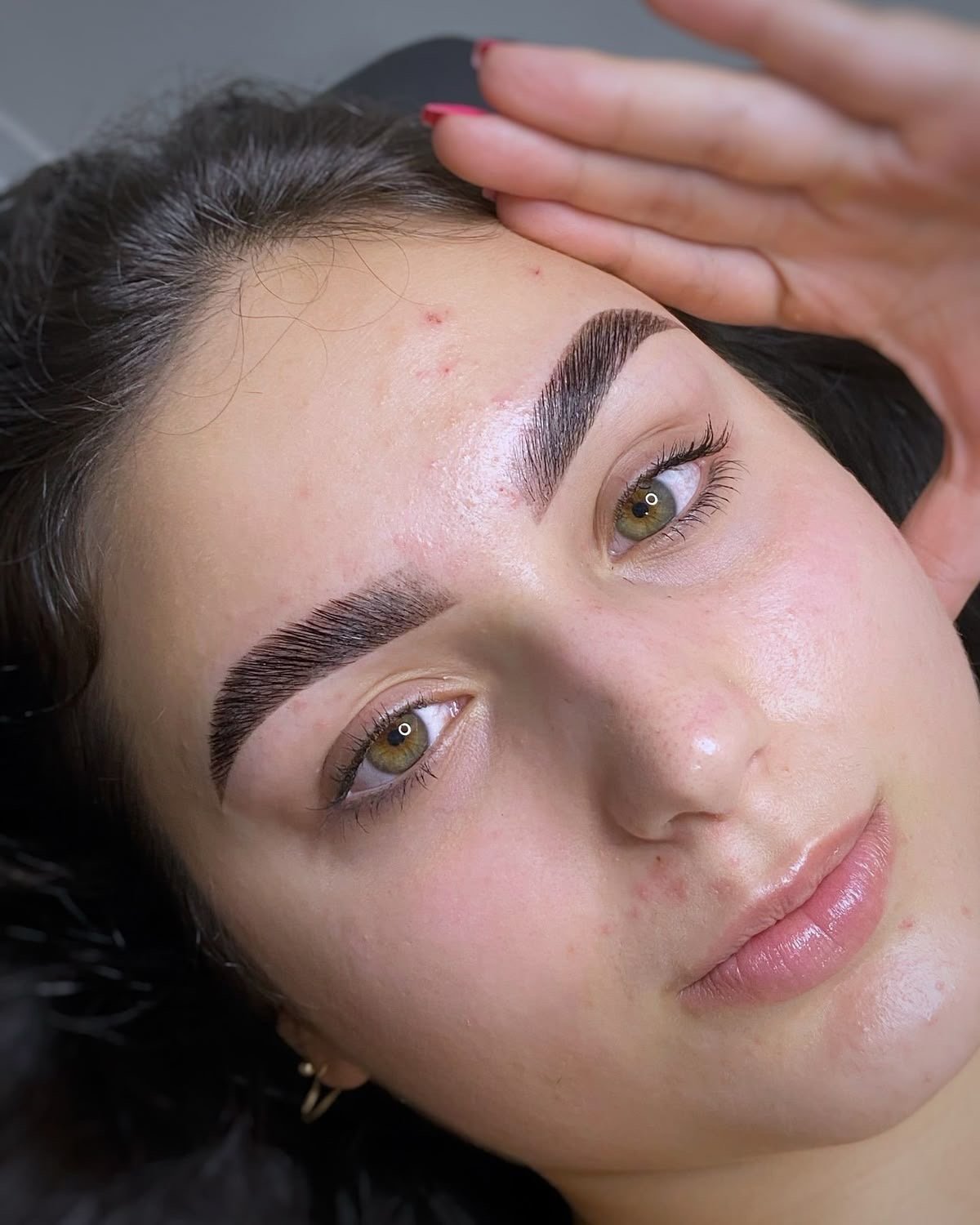 Mujer con cejas arregladas y piel brillante, recostada en una camilla con la mano en su frente.