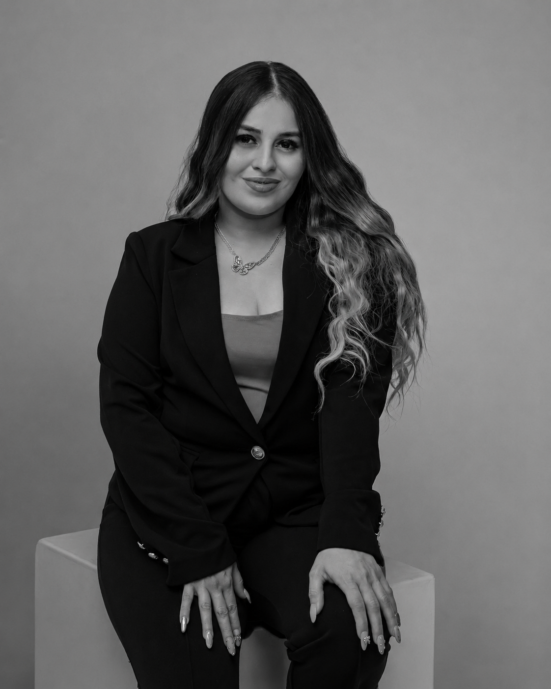 Retrato en blanco y negro de una mujer sentada sobre un bloque, con cabello rizado y vestimenta formal, sonriendo moderadamente.