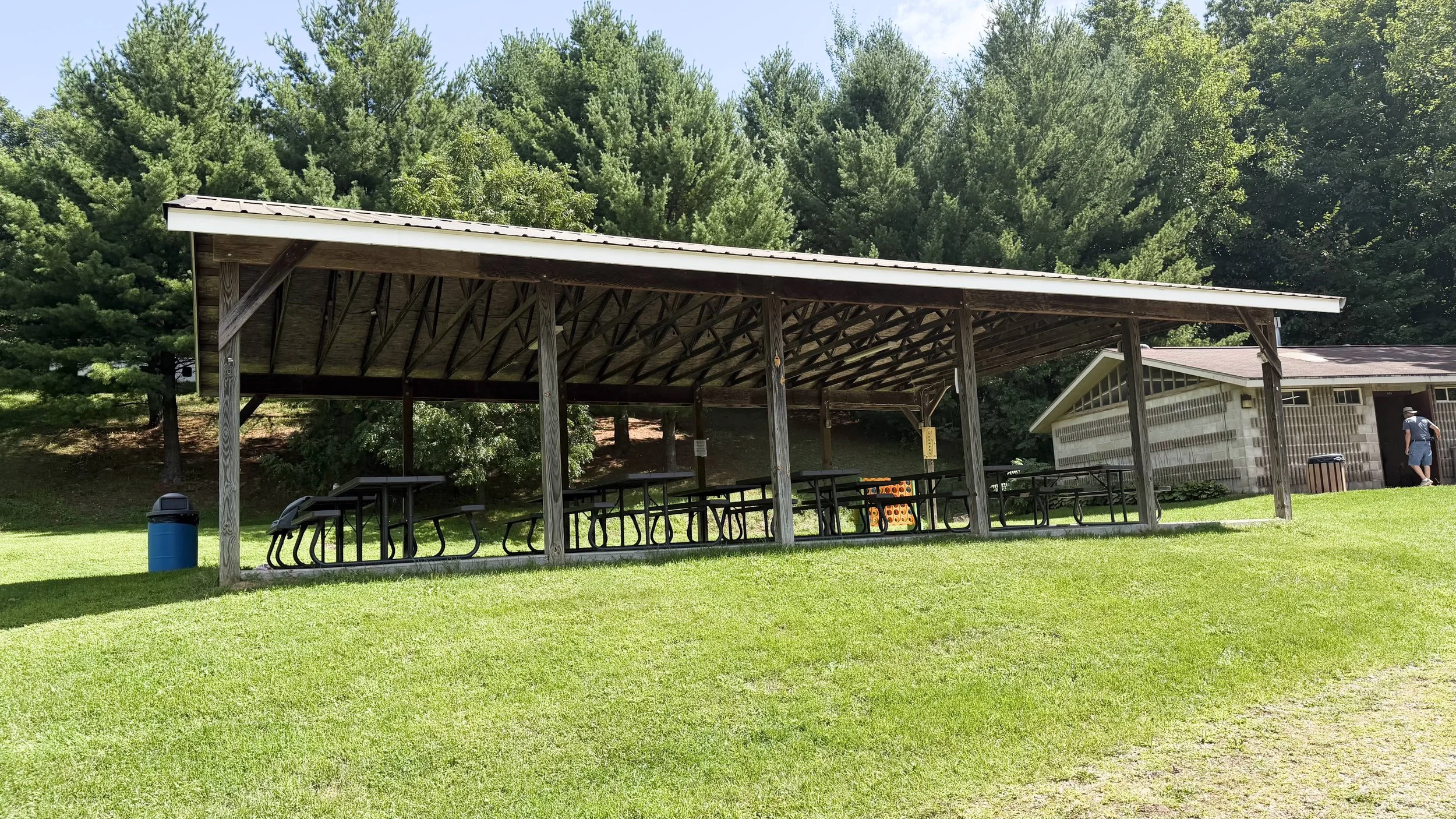 An outdoor park shelter with a sloped roof, supported by wooden posts, housing several picnic tables underneath. A blue trash can is near the shelter, and a person is walking near a small building in the background. The area is grassy with trees in the background.
