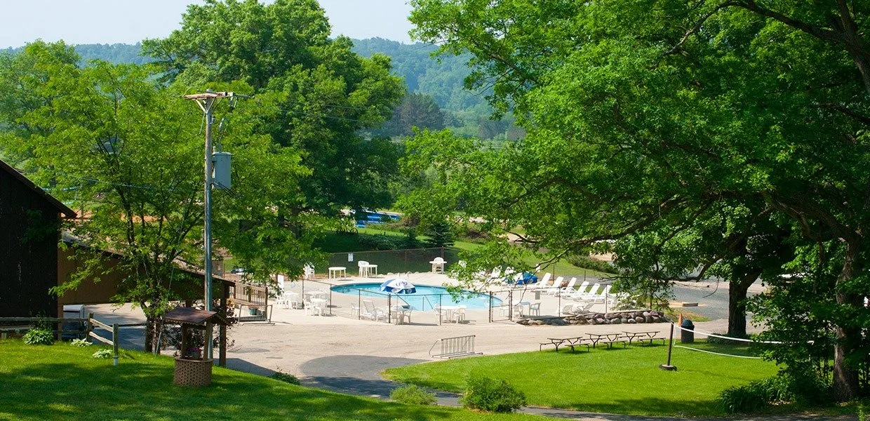 A swimming pool surrounded by lounge chairs, umbrellas, and trees in a outdoor recreational area.