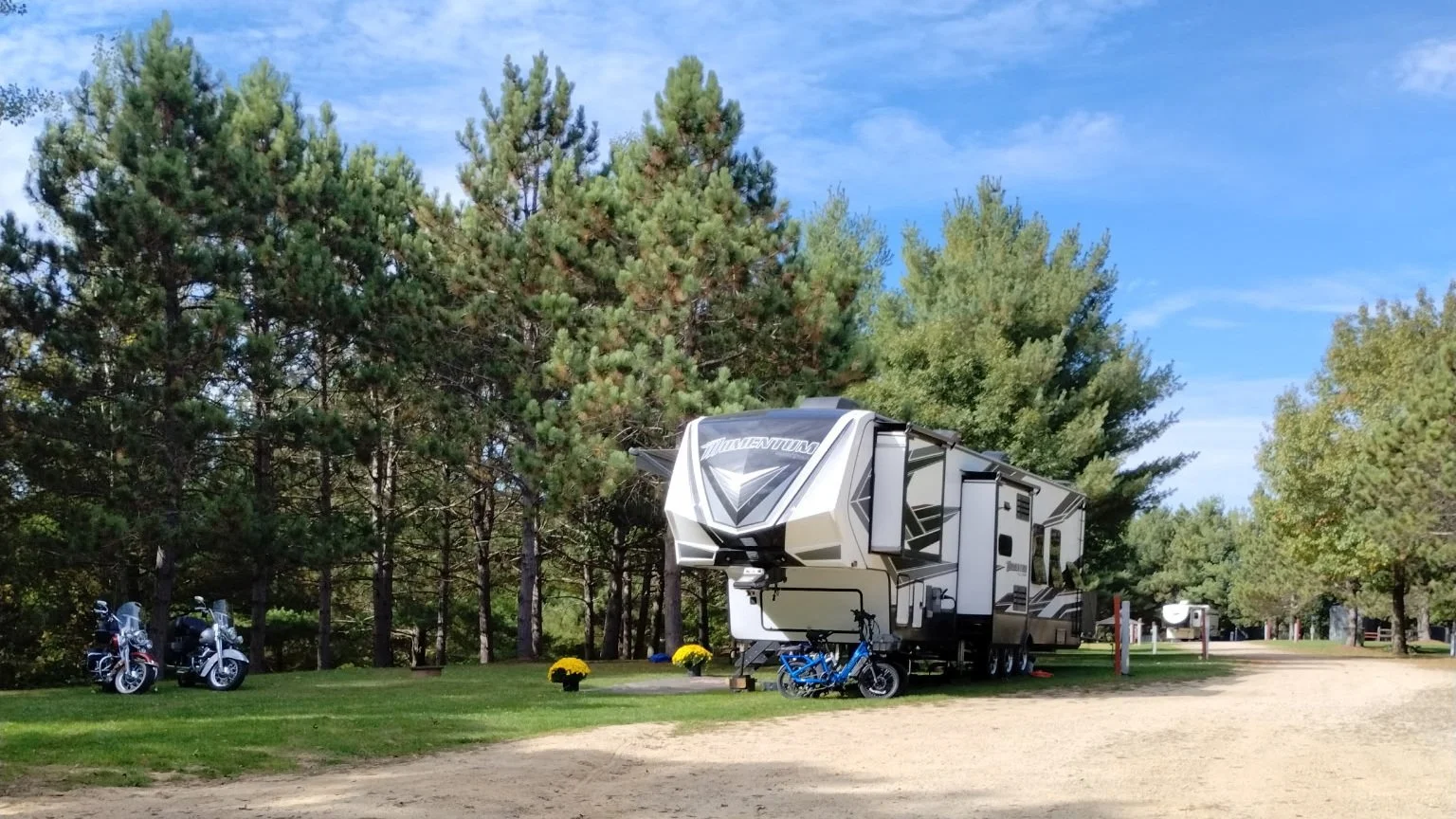 A white and black RV parked among green pine trees at a campsite with two motorcycles and a bicycle nearby, on a sunny day.