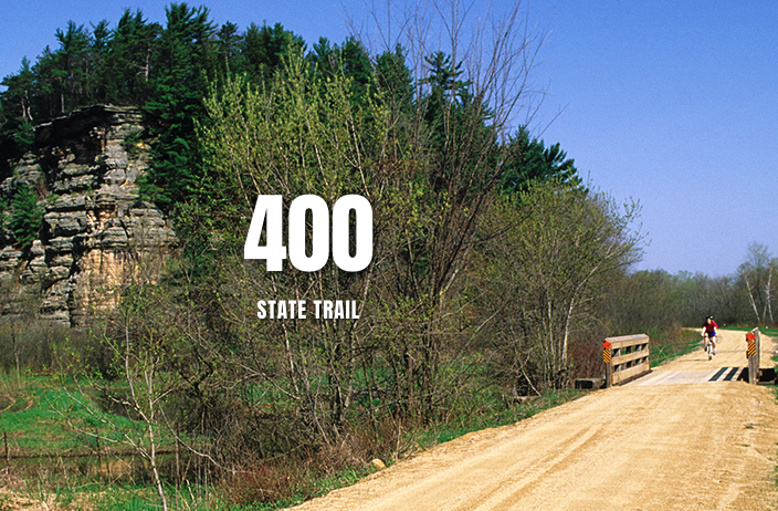 Dirt road crossing a small bridge with a cyclist and a pedestrian, surrounded by trees and a rocky cliff, with the text '400 STATE TRAIL' overlayed.