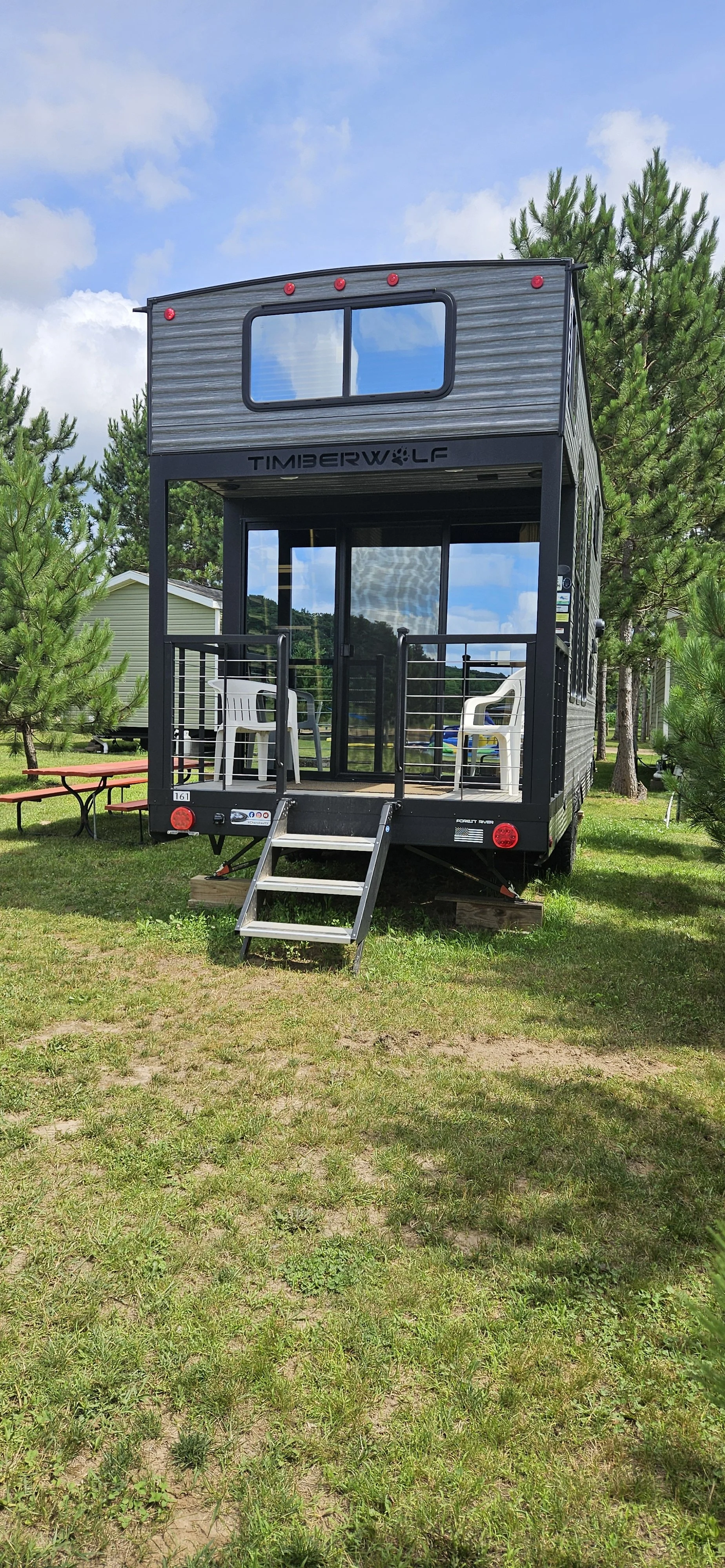 A black Timberwolf tiny home on wheels with a small front porch, two chairs inside, and surrounded by trees on a grassy area.