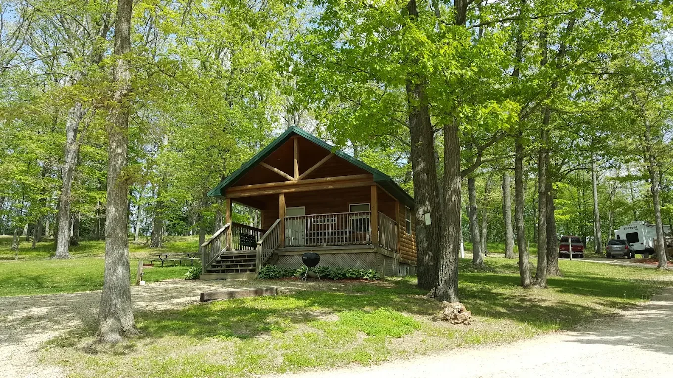 A small wooden cabin with a porch in a lush, green forested area, next to a gravel driveway with parked cars and an RV.