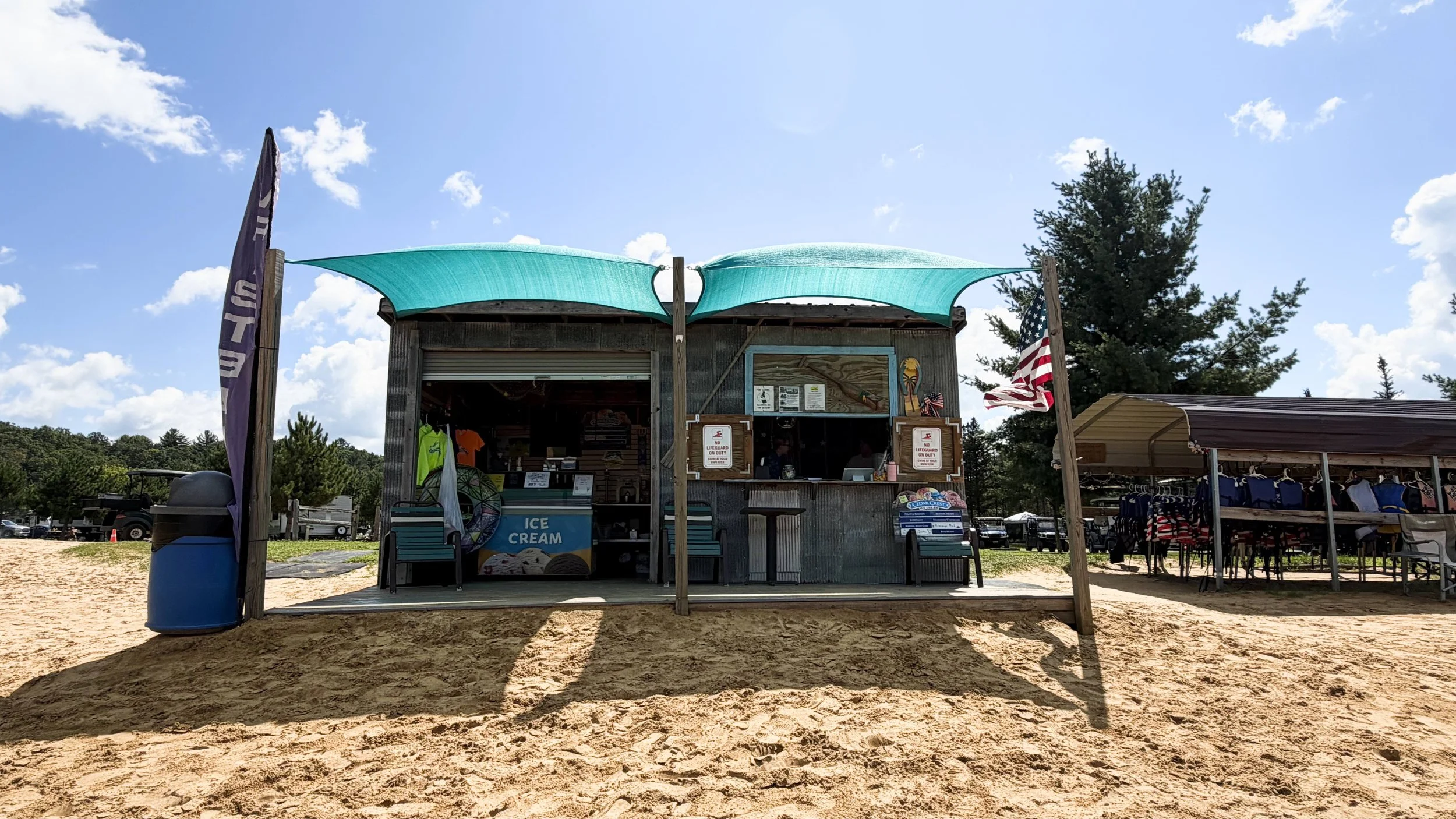 Beachside kiosk selling ice cream with an open window, American flags, and umbrellas, surrounded by trees and sandy ground under a partly cloudy sky.