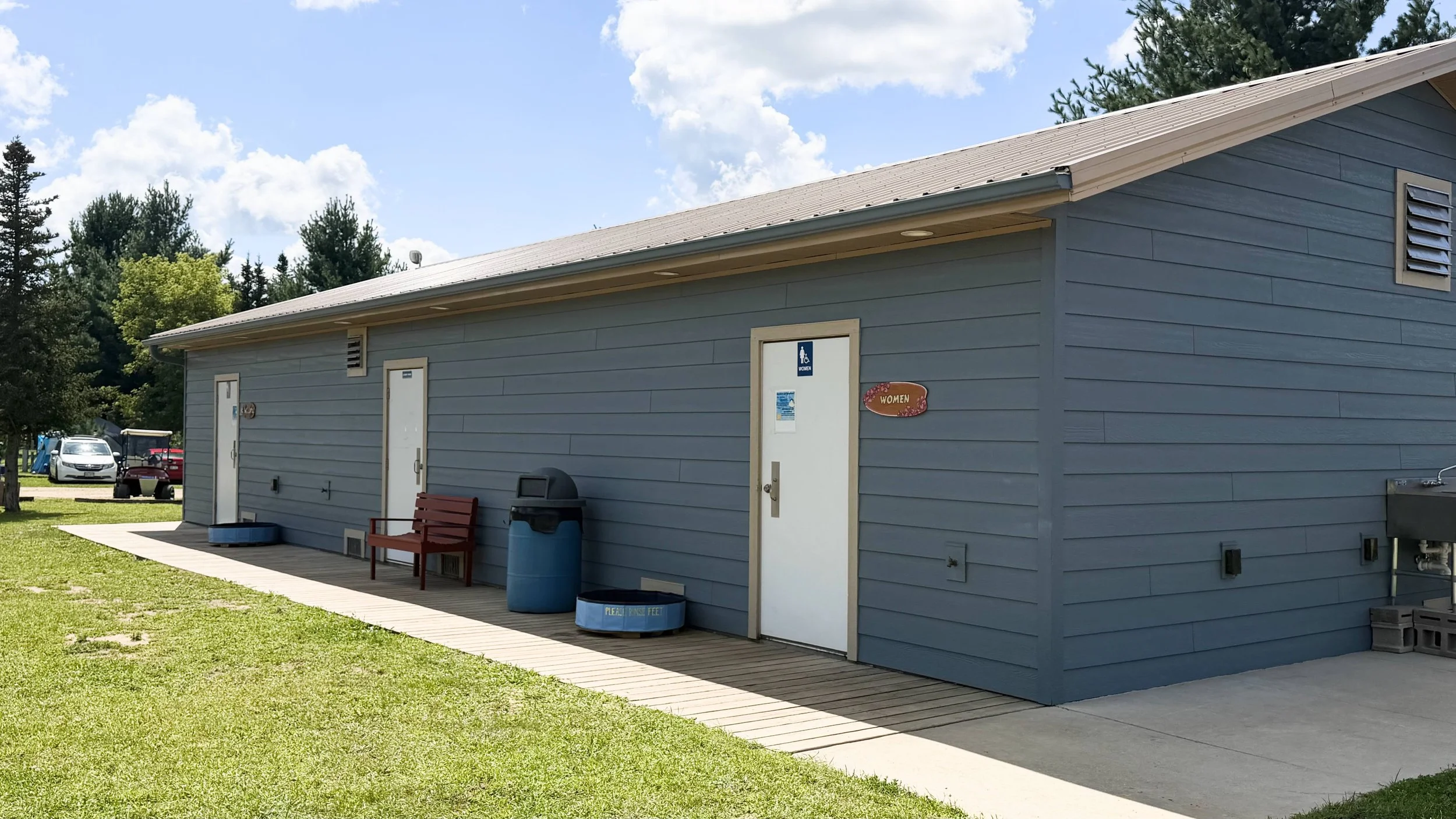 Blue public restroom building with two doors labeled for women and accessible, surrounded by green grass, a bench, trash can, and trees in the background under a partly cloudy sky.