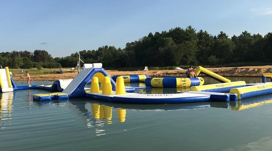 People enjoying on a large blue and yellow inflatable water obstacle course in a lake, with a wooded shoreline in the background.