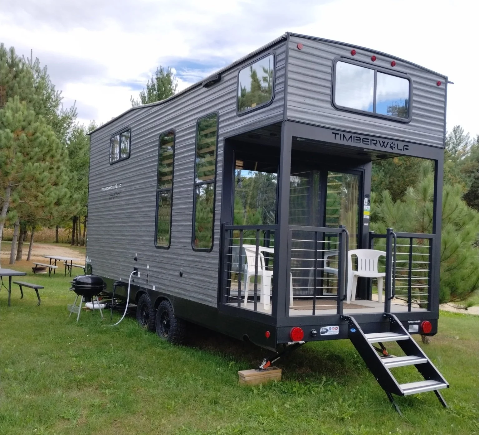 A modern tiny house on wheels with a small front porch, metal siding, and large windows, situated on a grassy area with trees in the background.