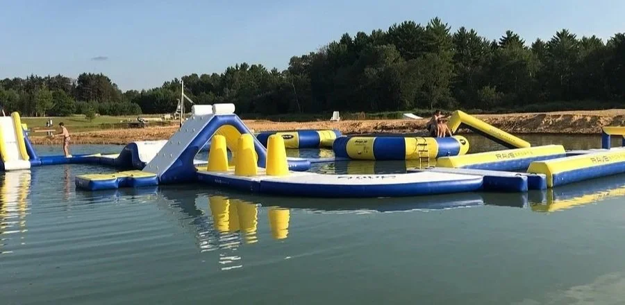 Inflatable water obstacle course on a lake with people playing and trees in the background.