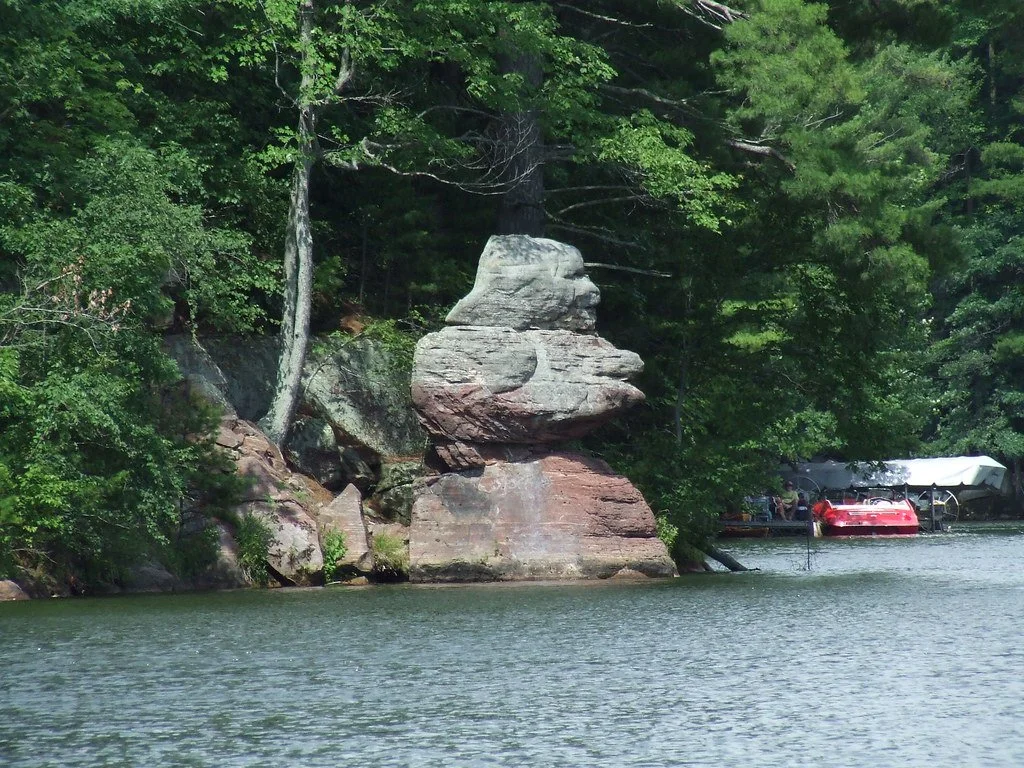 A lakeside scene featuring large rock formations and green trees, with a boat covered by a white tarp and a red boat docked at a marina in the background.