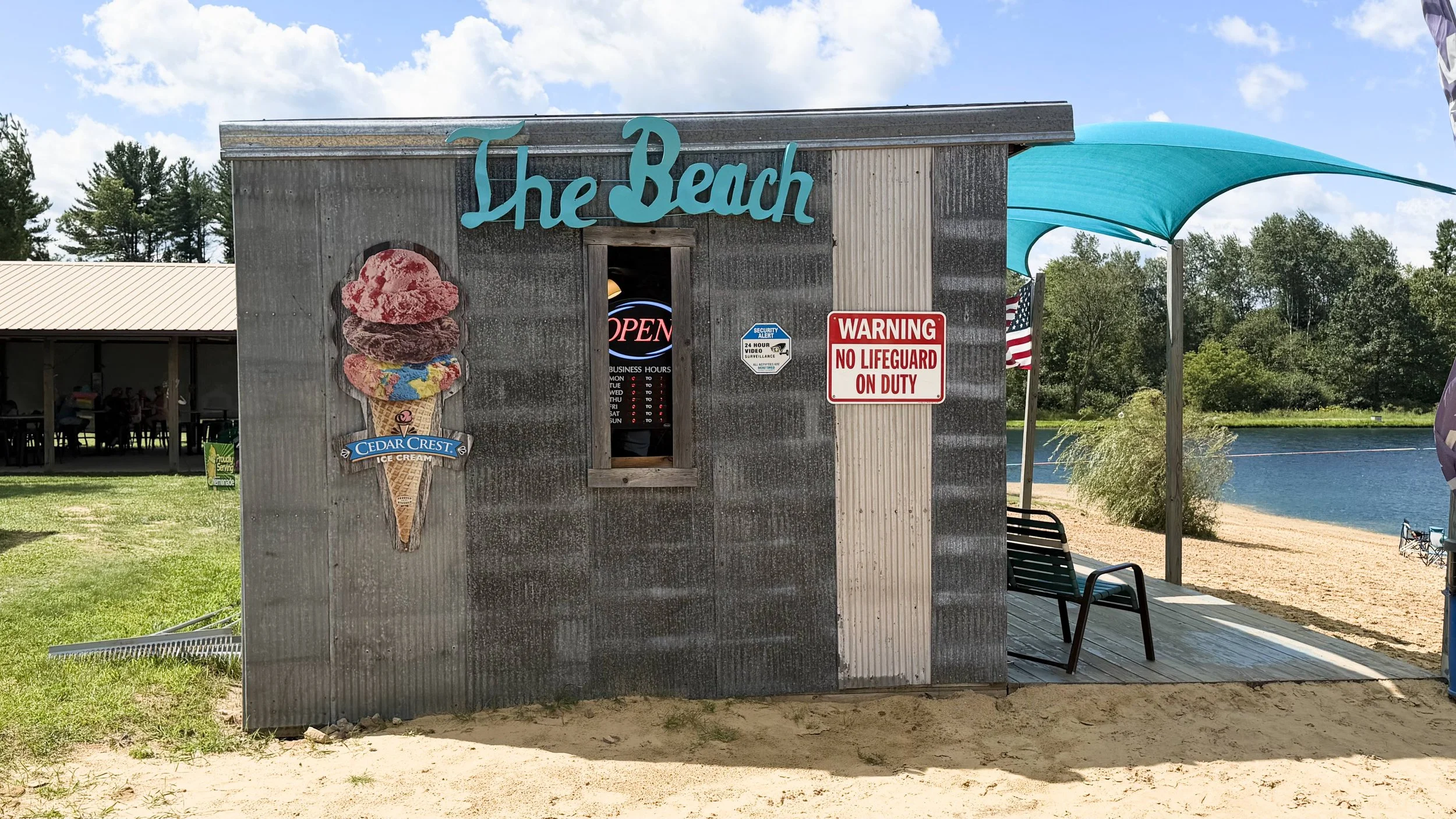 A small ice cream stand near a lake with a corrugated metal exterior, a large ice cream cone graphic, and a sign that says 'The Beach'. There is a blue sunshade, a red warning sign, and an American flag. Trees and water are in the background.