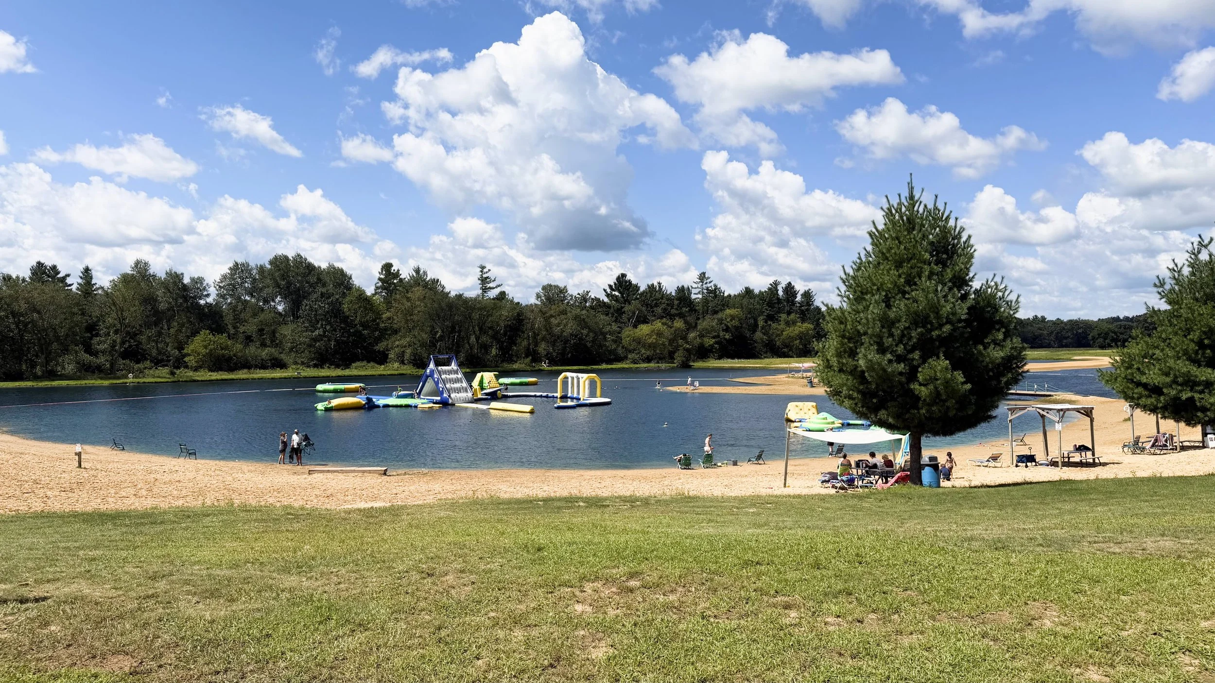 A lakeside scene with sandy beach, green grass, trees, and inflatable water park equipment in the lake, with people relaxing and enjoying the sunny day.