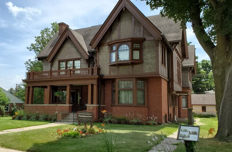 Historic two-story house with brick and wood siding, a front porch, and large windows, surrounded by a well-maintained lawn and trees.