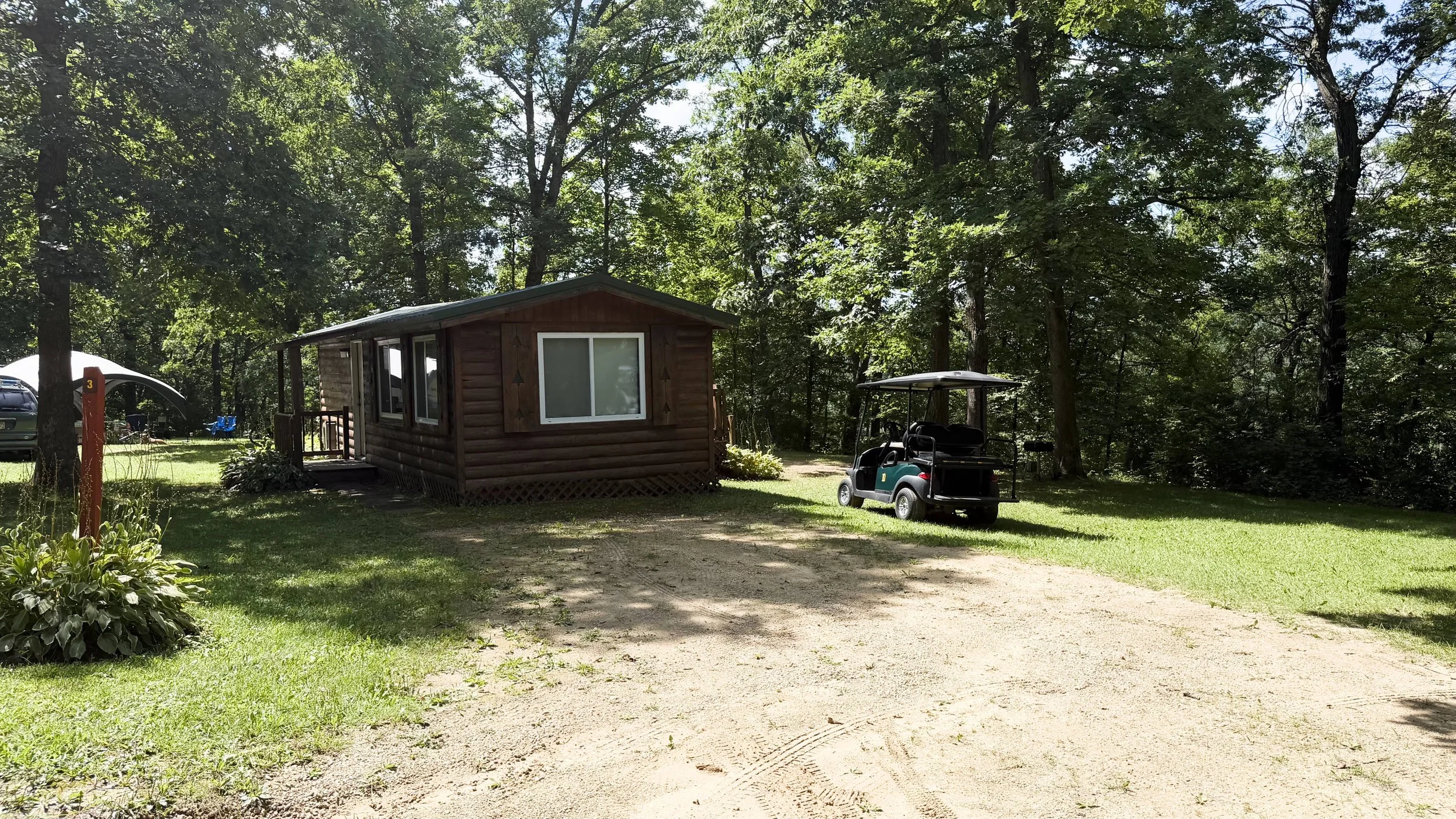 A small wooden cabin on a grassy area surrounded by trees, with a golf cart parked nearby and a few cars in the background.