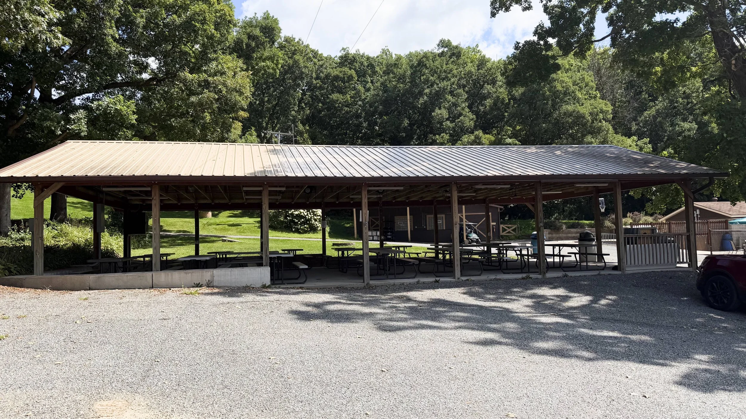 Empty outdoor pavilion with picnic tables, surrounded by trees and a gravel parking area.