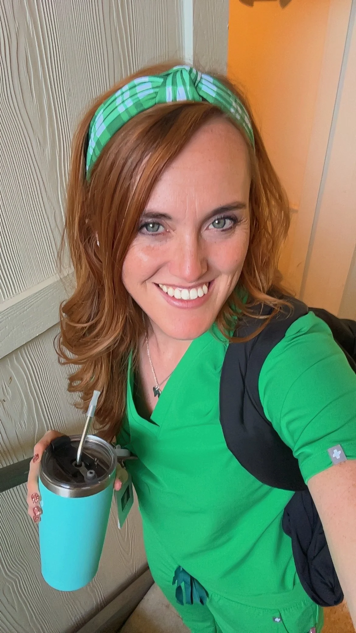 A woman with red hair wearing green scrubs and a green and white checkered headband, smiling and holding a blue insulated drinking cup with a straw.