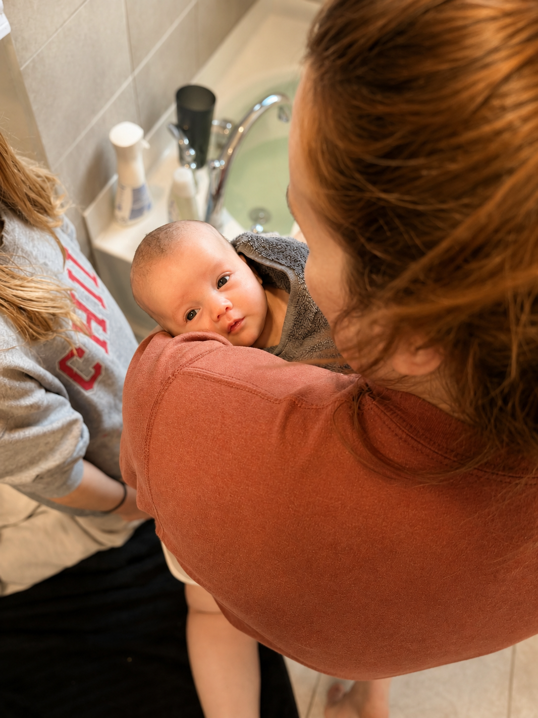 A woman holding a baby while standing over a sink, with another person partially visible nearby.