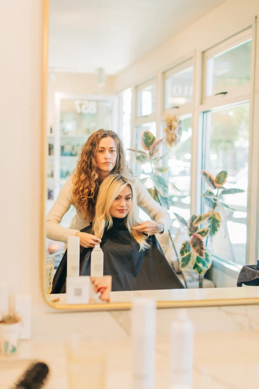 A woman getting her hair styled at a hair salon, with mirrors, plants, and hair products visible in the background.