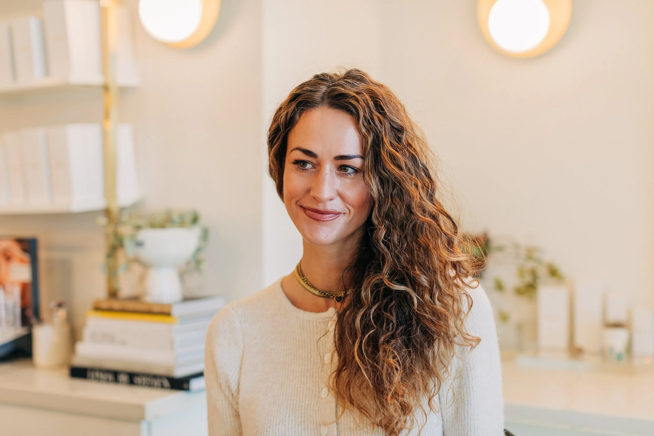 A woman with long, curly, reddish-brown hair smiling in a well-lit room with bookshelves in the background.