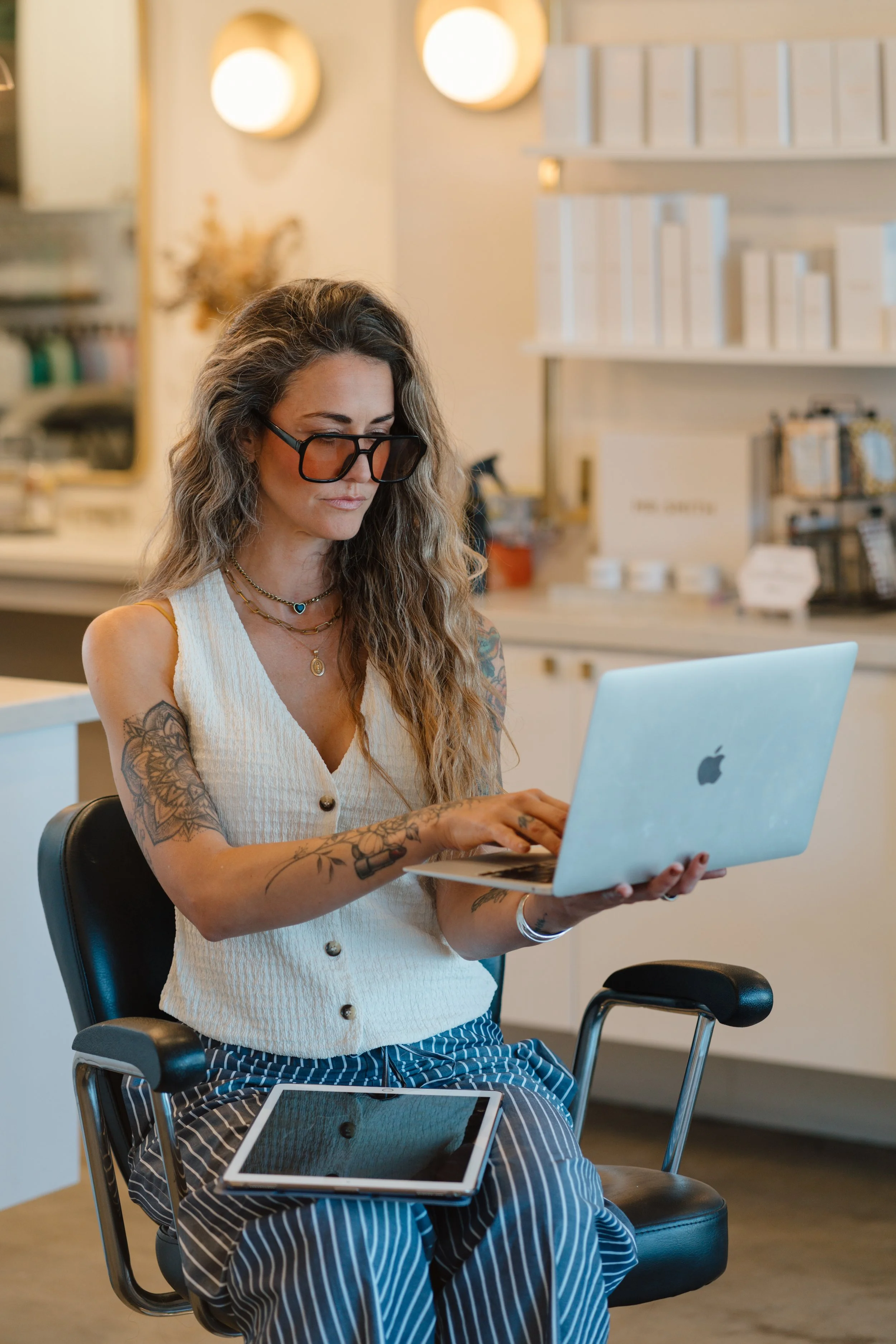 A woman with glasses and tattoos wearing a sleeveless top and striped skirt, sitting in an office chair, looking at a silver laptop. There are digital devices on her lap and in front of her. The background includes shelves with books and circular wall decorations.