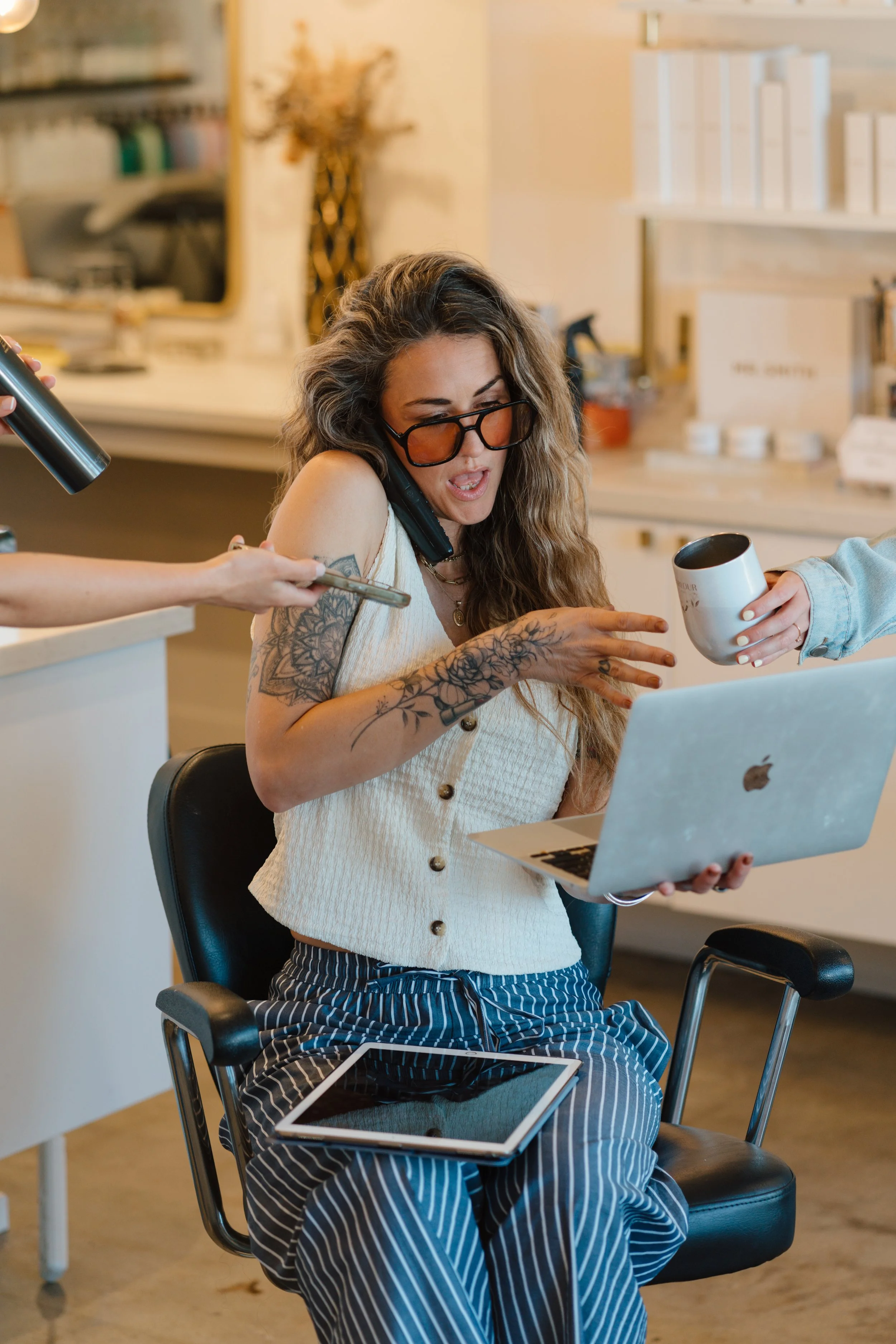 A woman with long curly hair and tattoos on her arms is sitting in a black office chair, wearing glasses, a sleeveless top, and striped pants. She is holding a notebook on her lap and is being handed a mug while talking on a cell phone.
