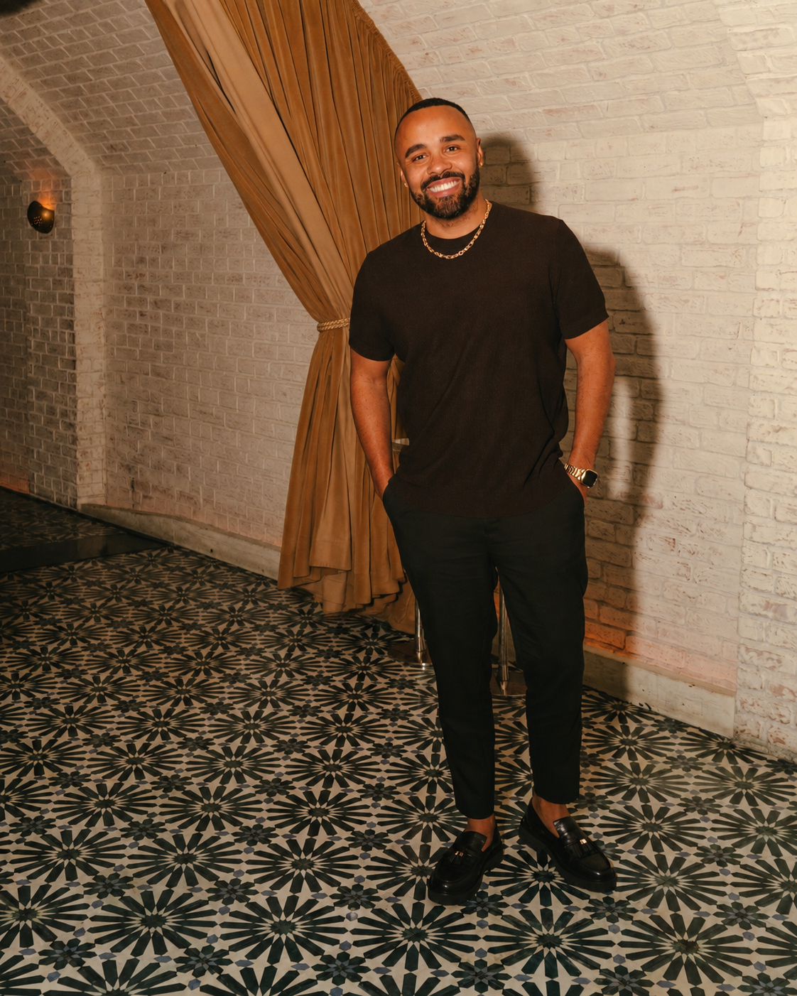 Chase Michael smiling in a black outfit against a white brick wall with draped curtains