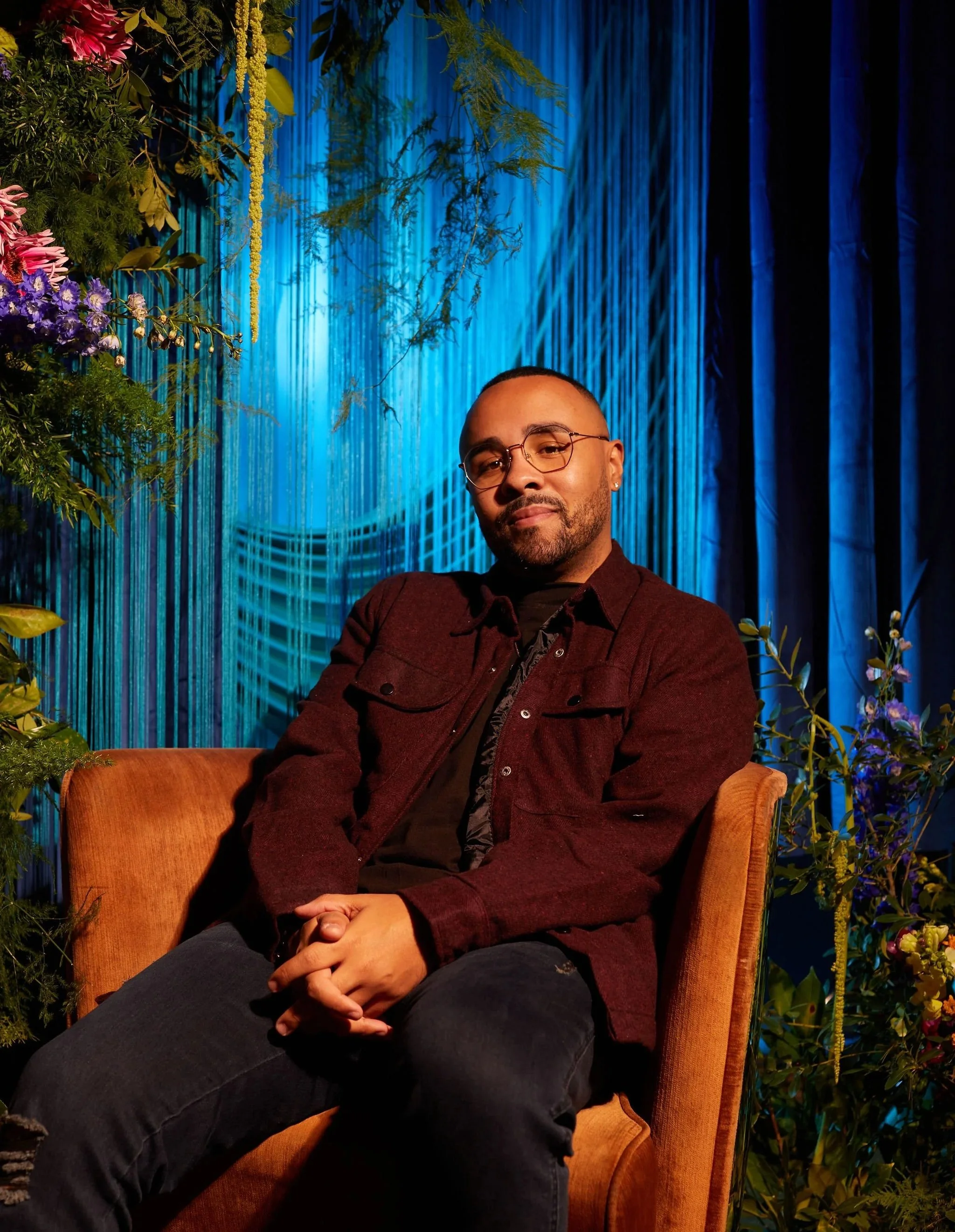 Chase Michael in glasses and a burgundy jacket, seated in an orange armchair against a blue-lit backdrop with flowers
