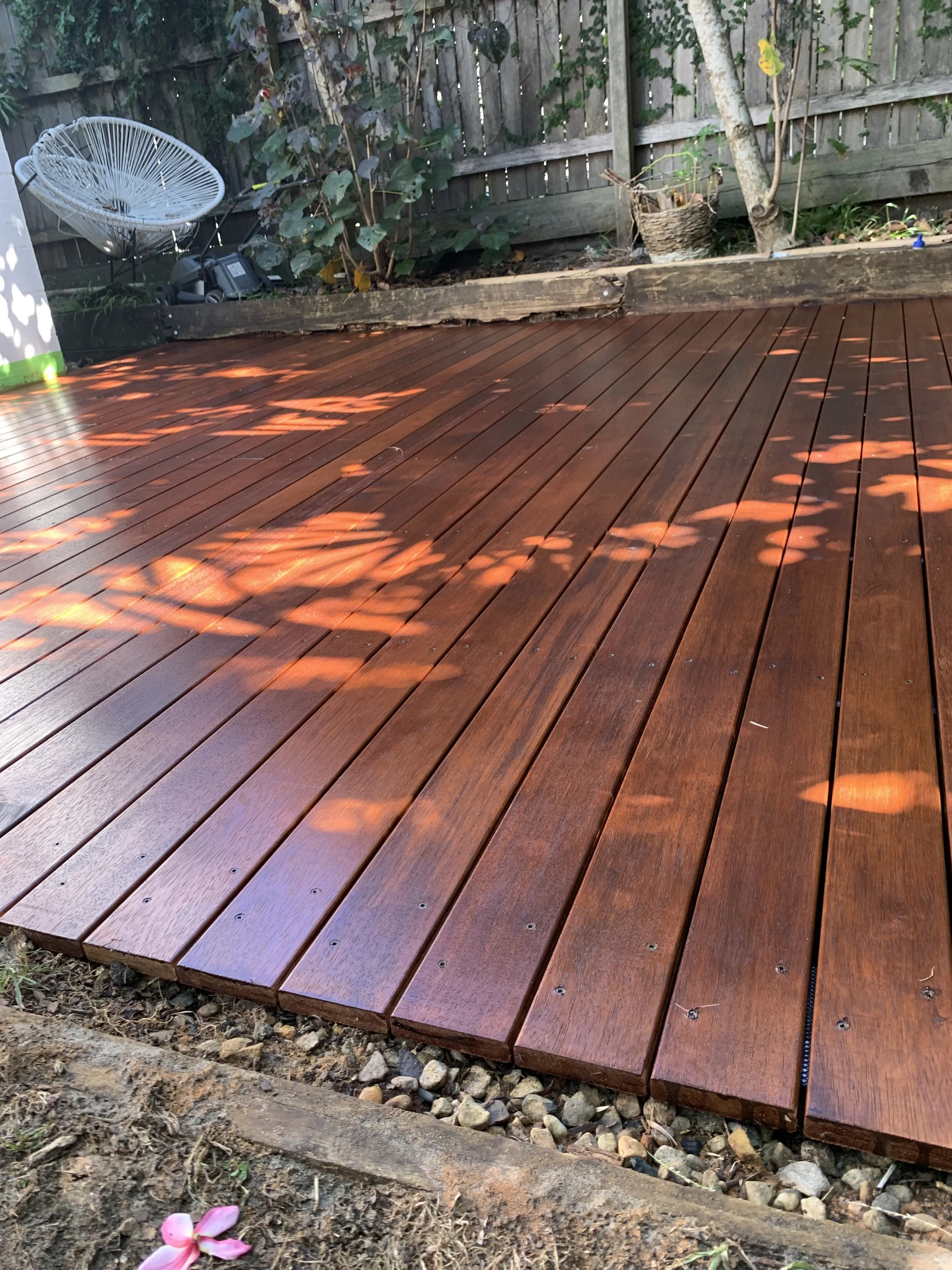 Freshly installed wooden deck in a backyard garden with green shrubbery and a wooden fence in the background; there are shadows of leaves on the deck, and some small pink flowers are visible at the lower left corner.