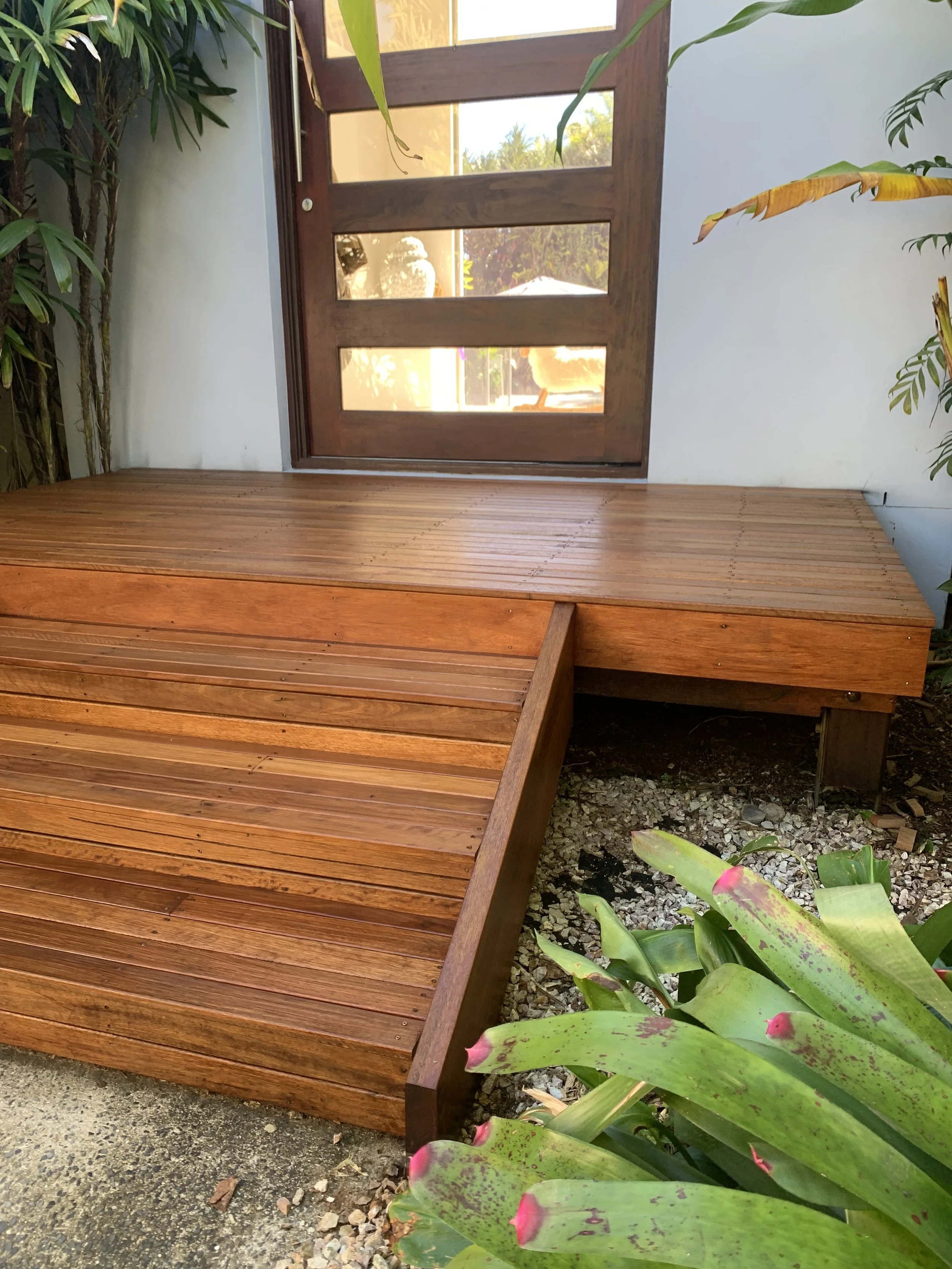 A wooden deck with a raised platform next to a glass-paneled door, surrounded by green plants with large leaves.