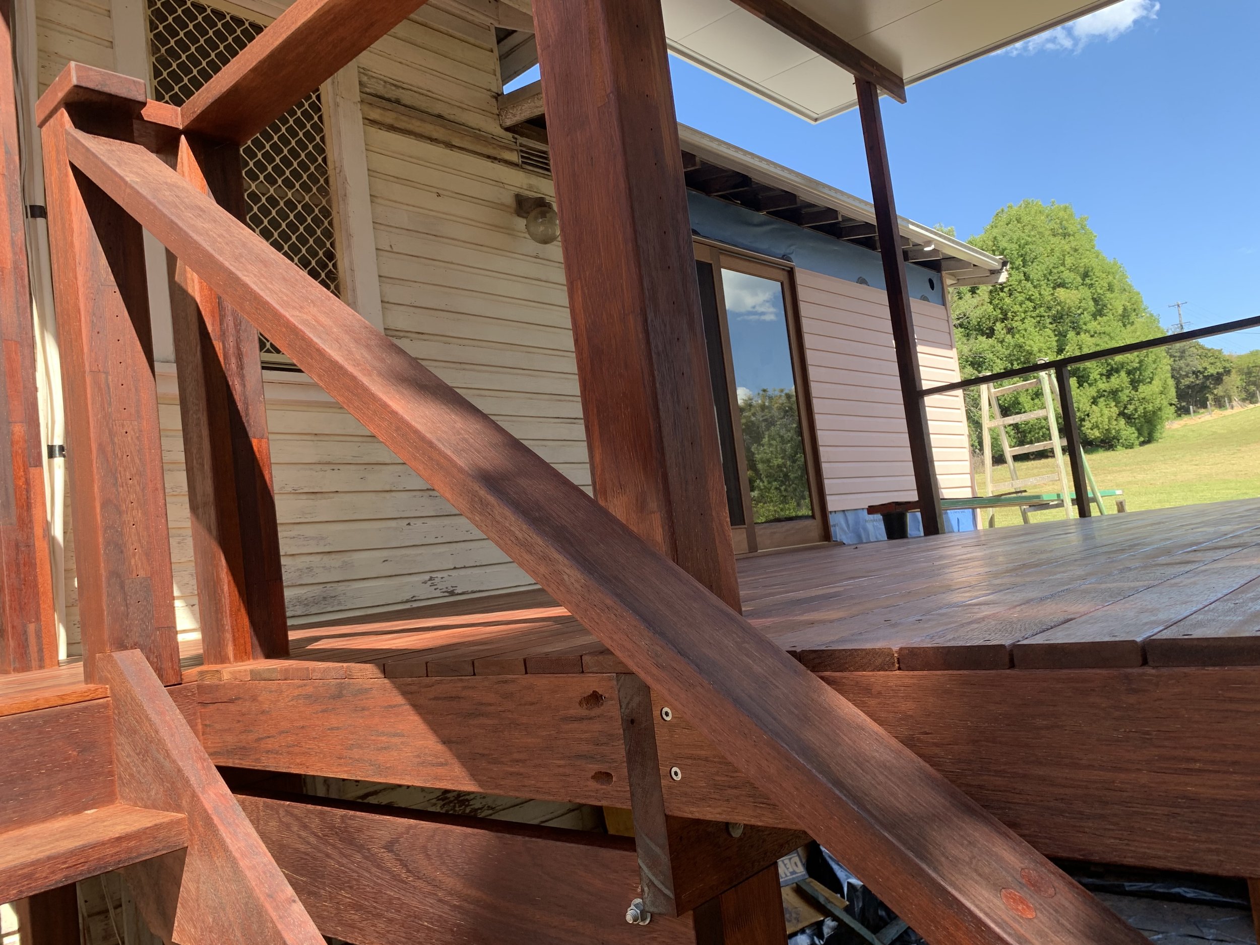 A wooden deck with stairs attached to the side of a house, with greenery and blue sky in the background.