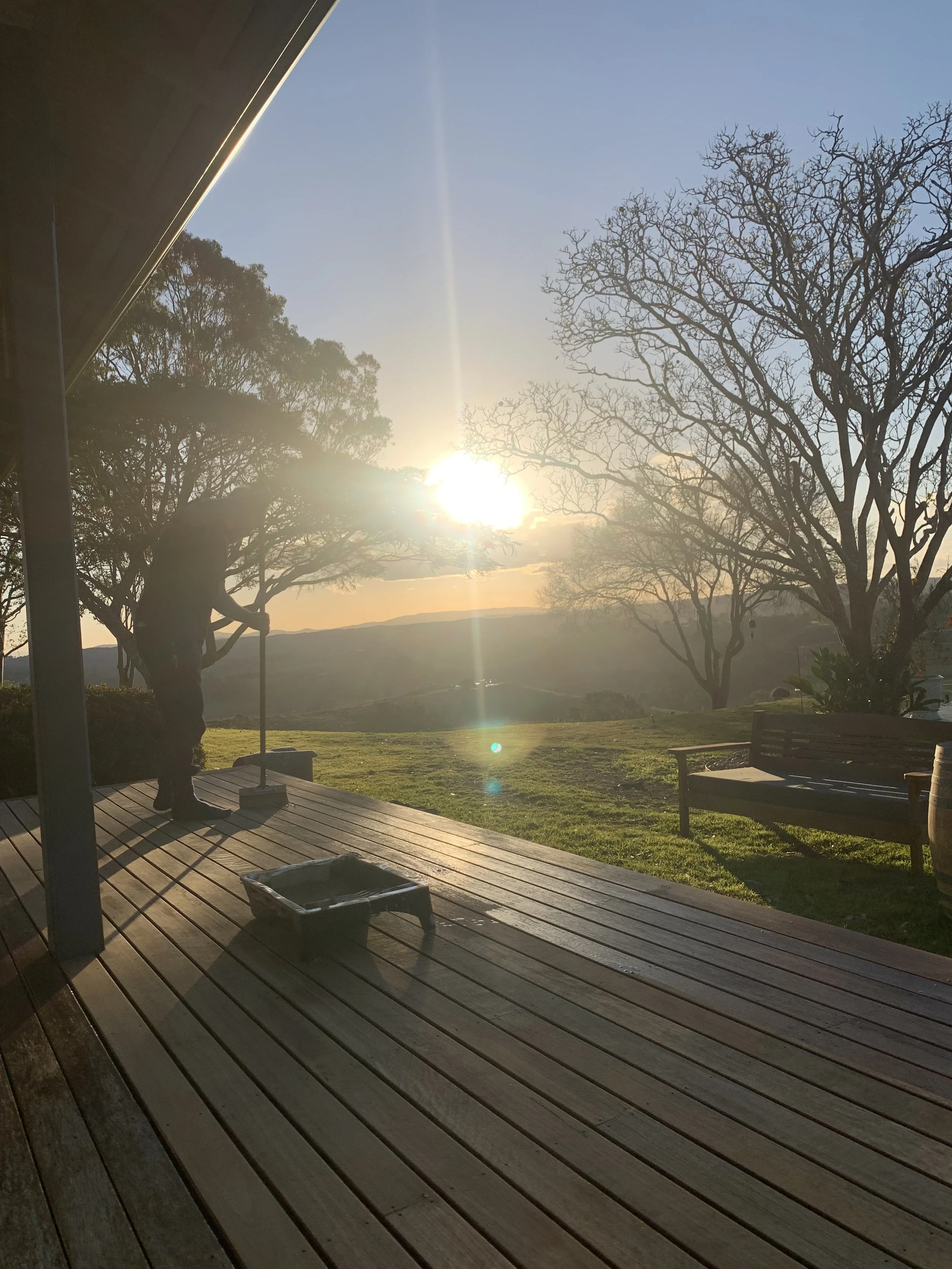 Sun setting over a clear sky with trees, seen from a wooden porch with benches and a small cart.