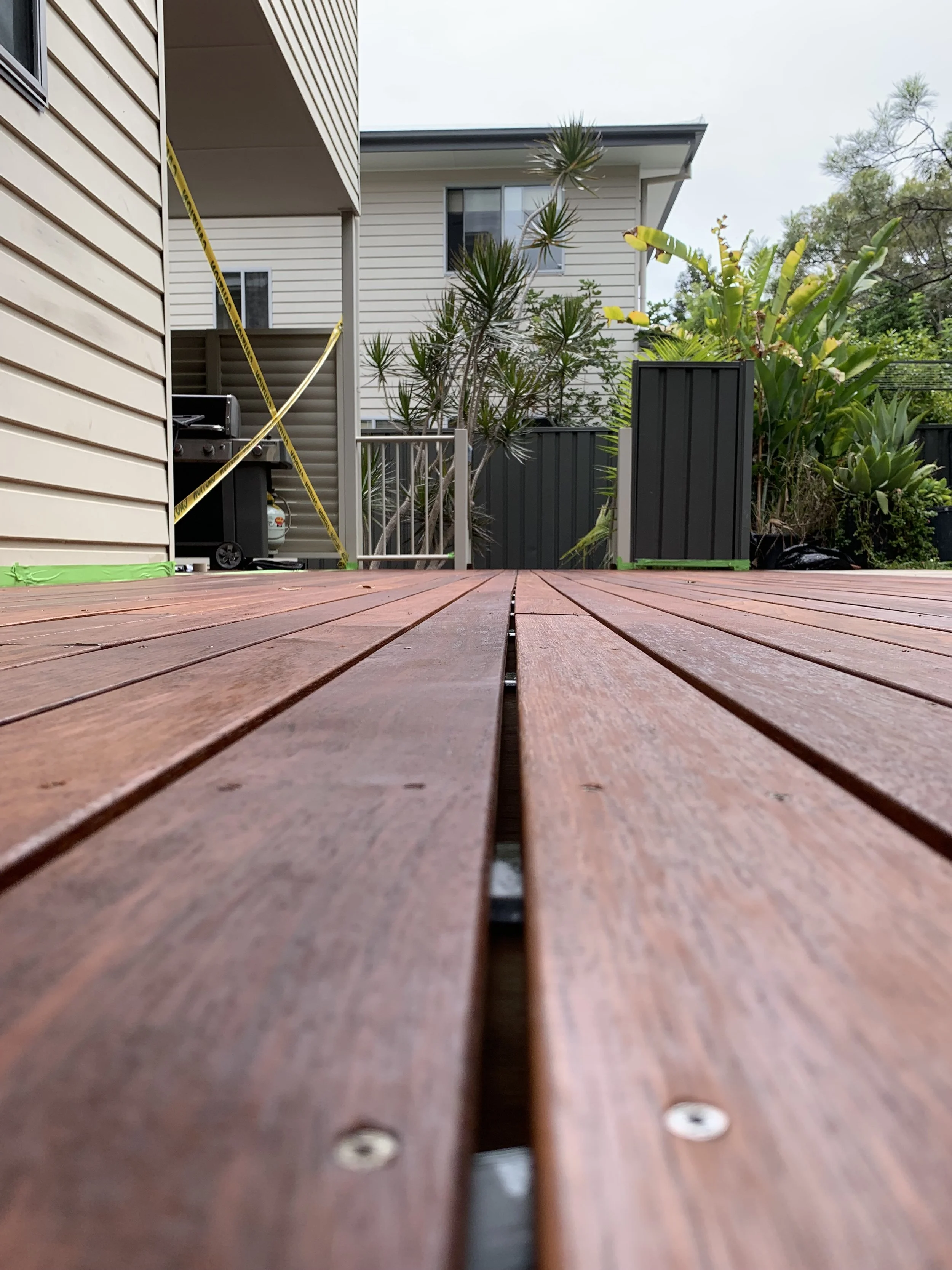 View of an outdoor wooden deck with plants in large planters, neighboring houses, and a gas grill.