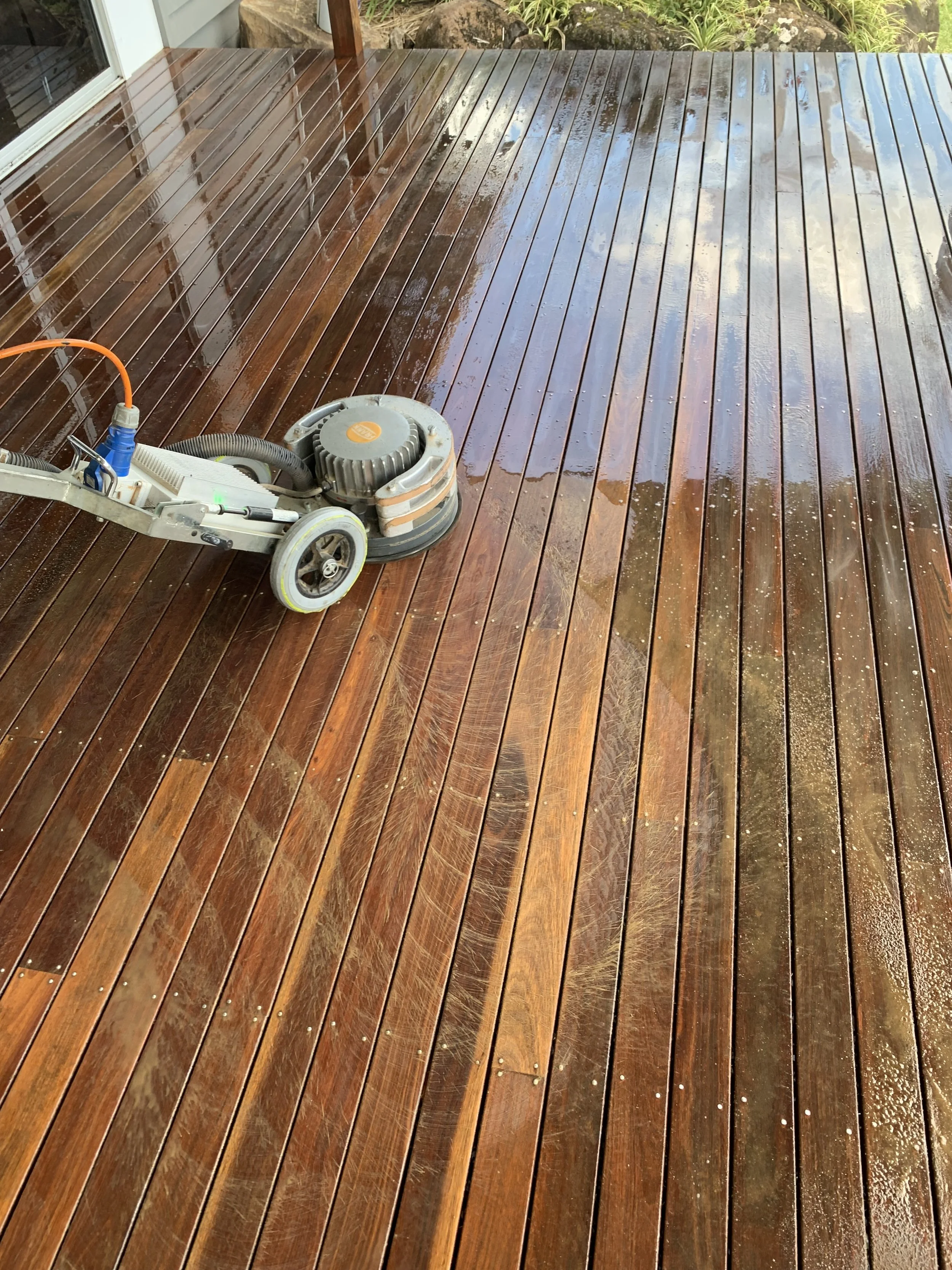 Wet stained wooden deck with a floor sanding machine in the lower left corner, some greenery at the back.