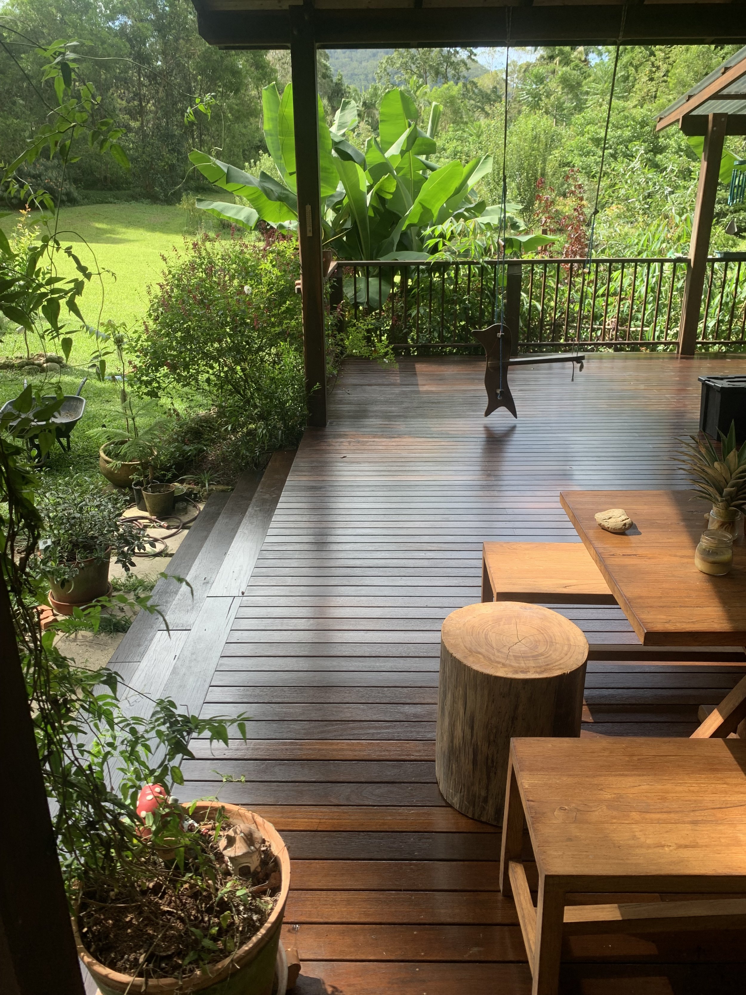 A wooden deck with a potted plant, a rustic wooden stool, and a wooden table with small items on it. Greenery and trees are visible beyond the deck.