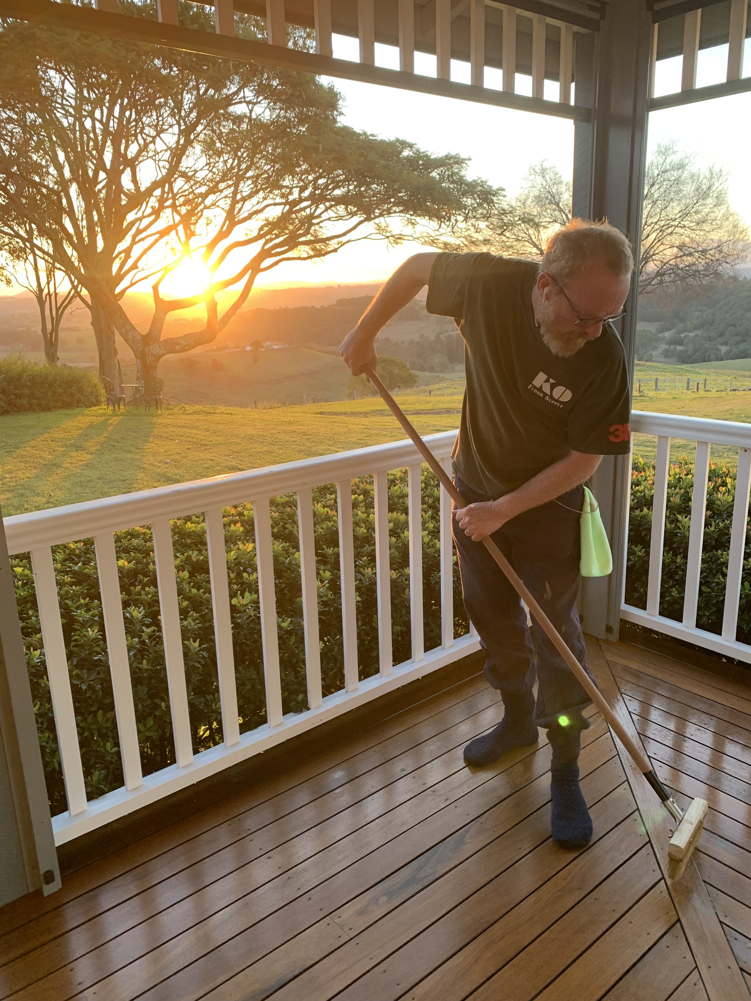 An older man with glasses and a beard is sweeping a wooden porch with a broom. The porch has white railing and is surrounded by lush greenery and trees. The sun is setting in the background, creating a warm glow.
