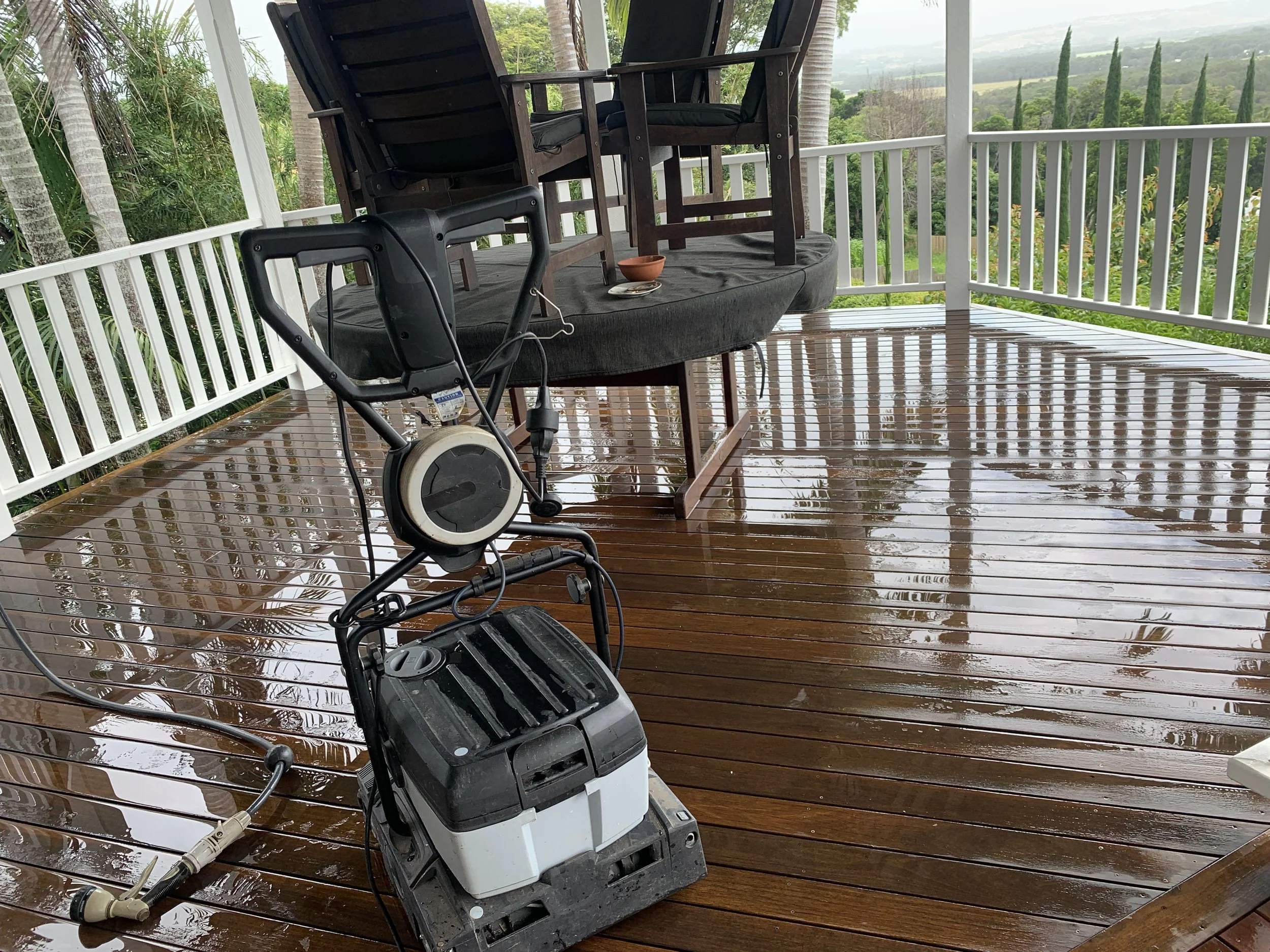 A wet wooden porch with outdoor furniture including three brown chairs with black cushions, a small table with a ceramic bowl, and a standing device with a cable. The background features lush green trees and rolling hills.