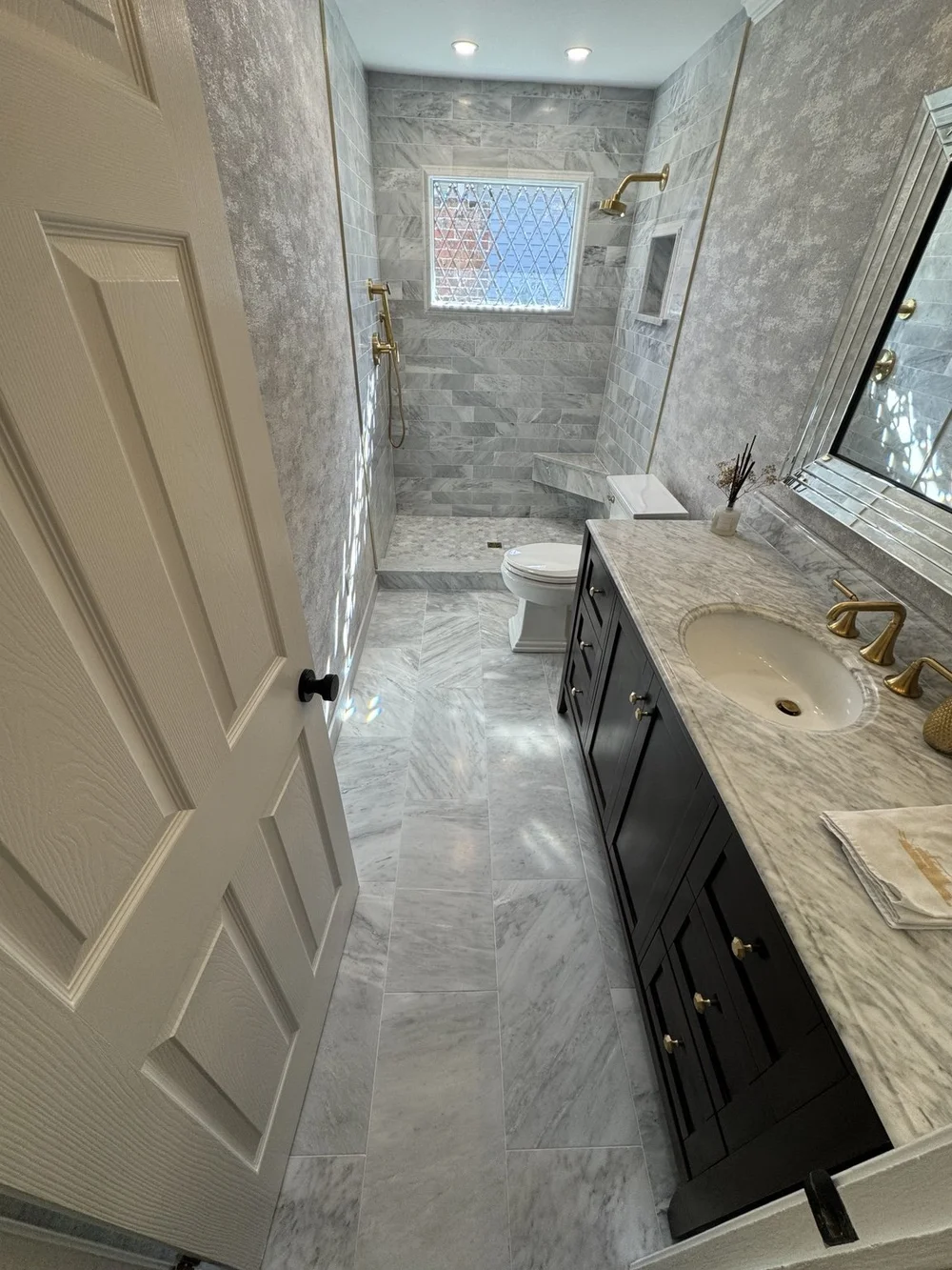 Complete bathroom remodel with custom shower, marble-look tile flooring, vanity, countertop, and brass fixtures.