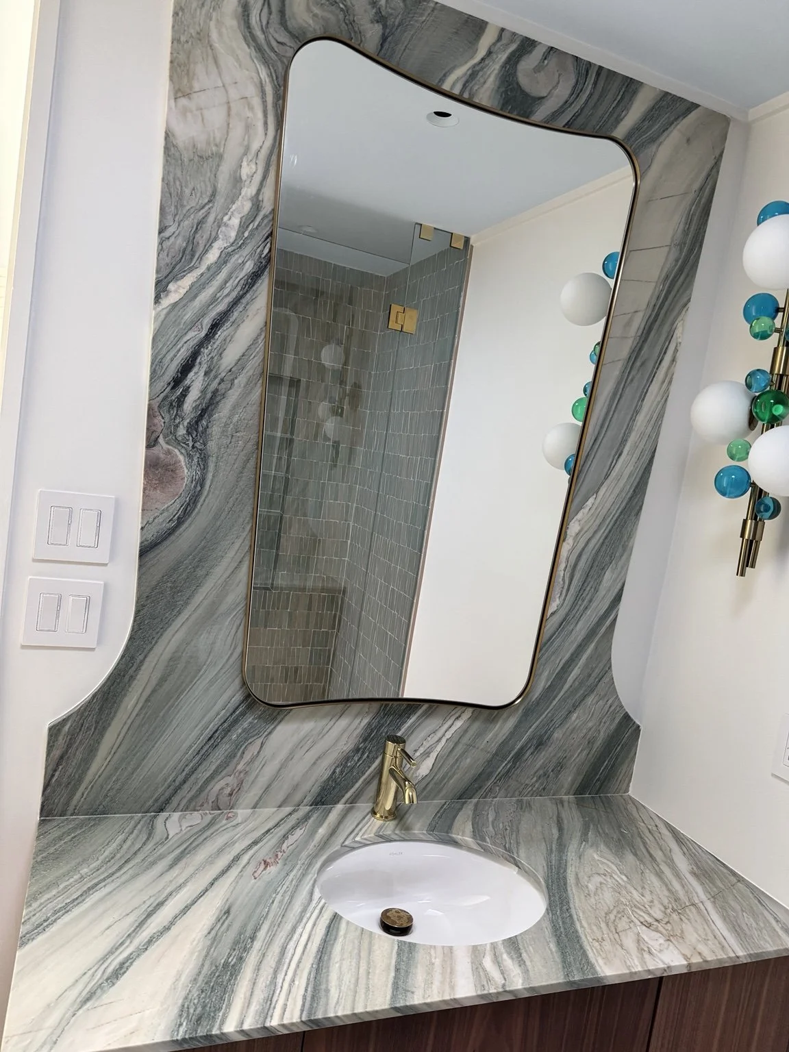 Veined vanity counter and full-height feature wall with a gold faucet, sculptural mirror, and colorful globe sconces by EC Construction LLC.
