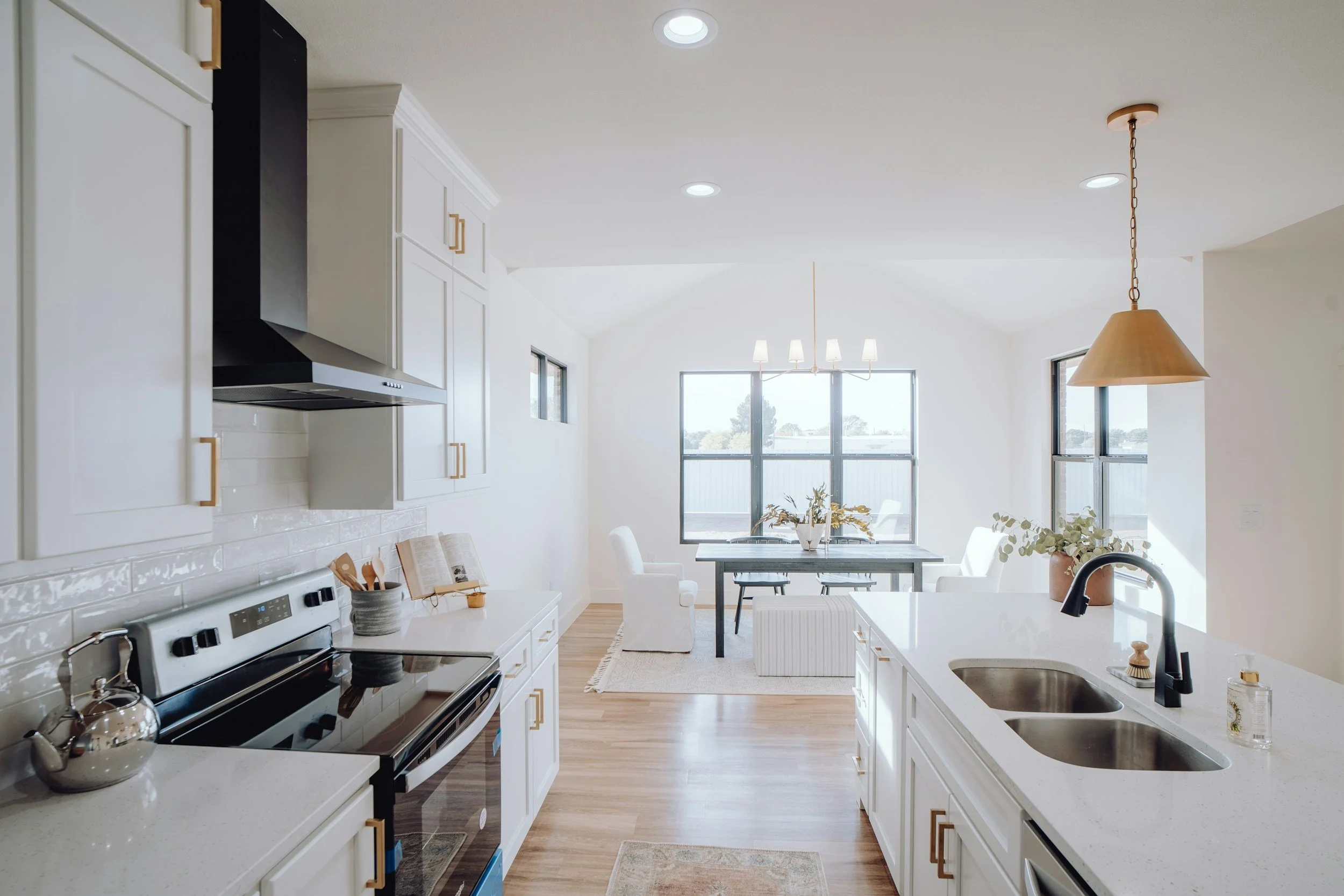 A bright, modern kitchen with white cabinetry, a black stove, white countertops, and a double sink. The kitchen opens into a dining area with a table, chairs, and large windows letting in natural light. A hanging pendant light and a chandelier are visible.