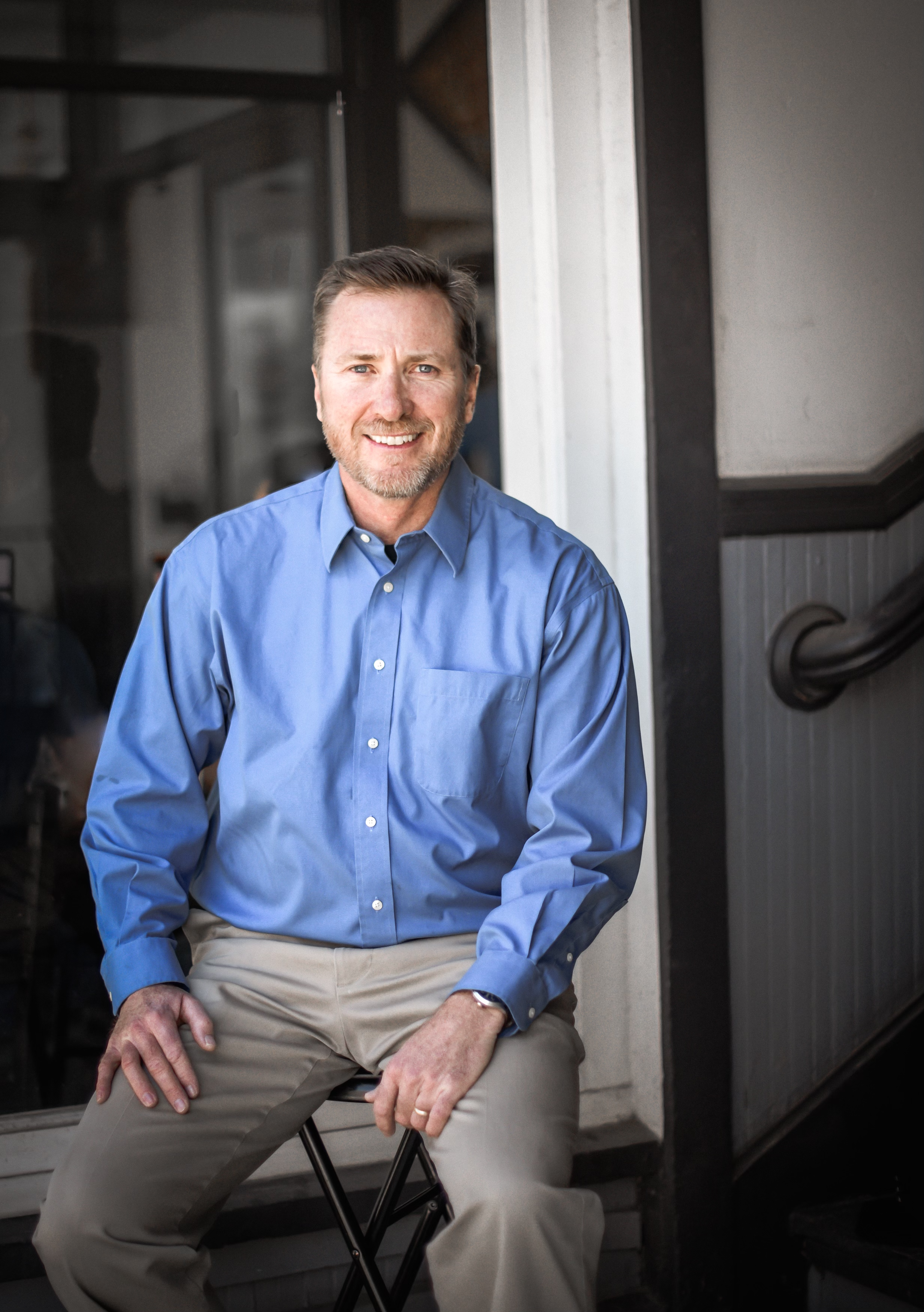A smiling middle-aged man with a beard, wearing a blue long-sleeve button-up shirt and beige pants, sitting on a black folding chair in front of a gray and black construction or industrial background.