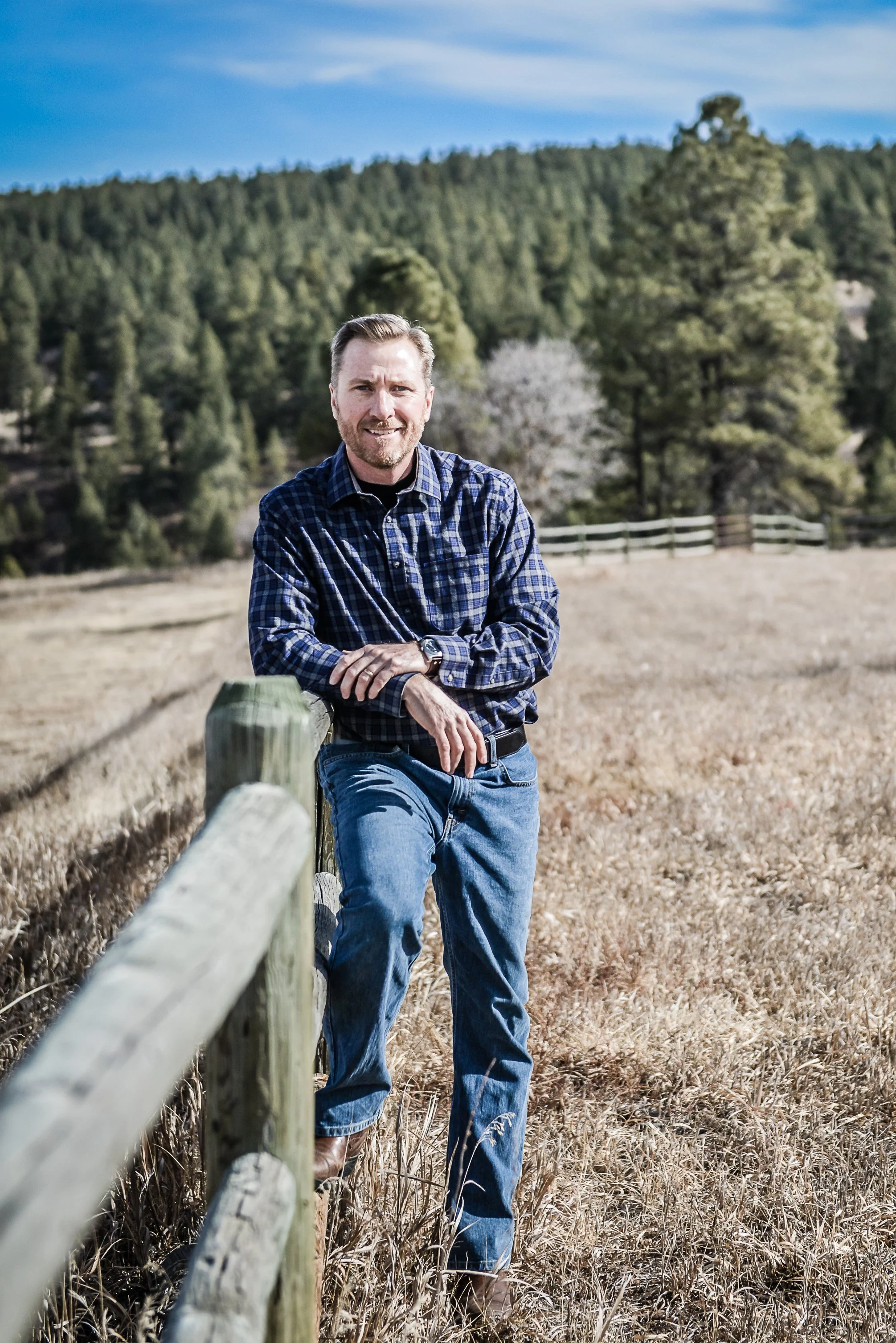 A man with a beard in a blue plaid shirt and jeans leans against a wooden fence on a grassy field with trees and a forested hillside in the background.