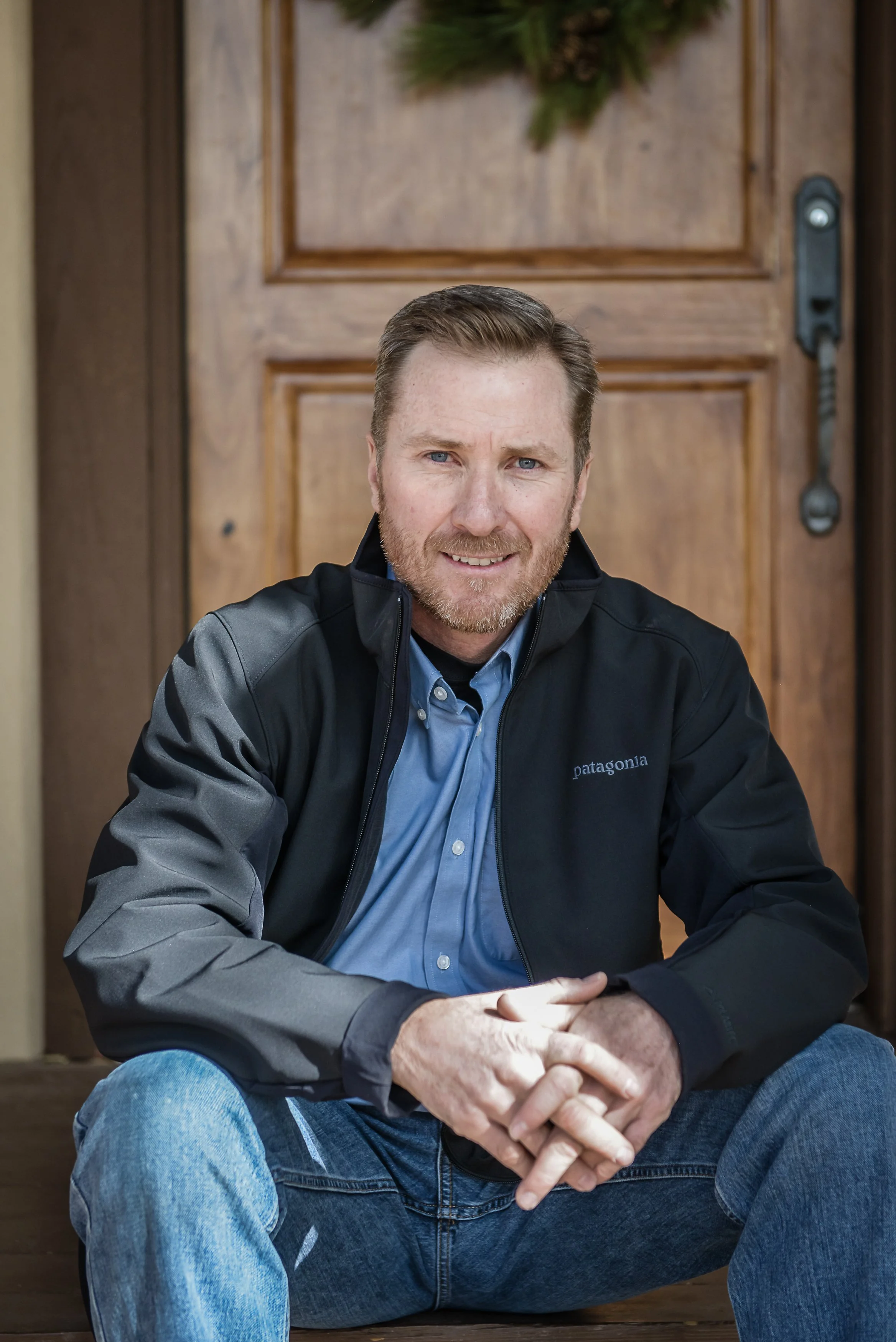 A man with short brown hair and a beard, wearing a black Patagonia jacket and blue shirt, sitting in front of a wooden door with a wreath on it.
