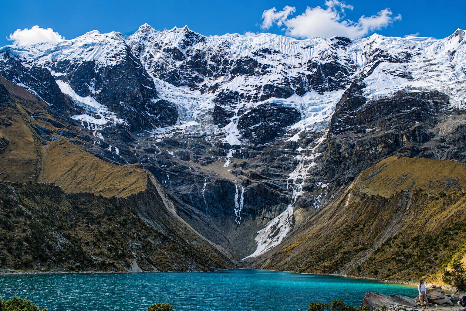 Snow-capped mountains surrounding a glacial lake with turquoise water, rocks, and a person standing on the shore.