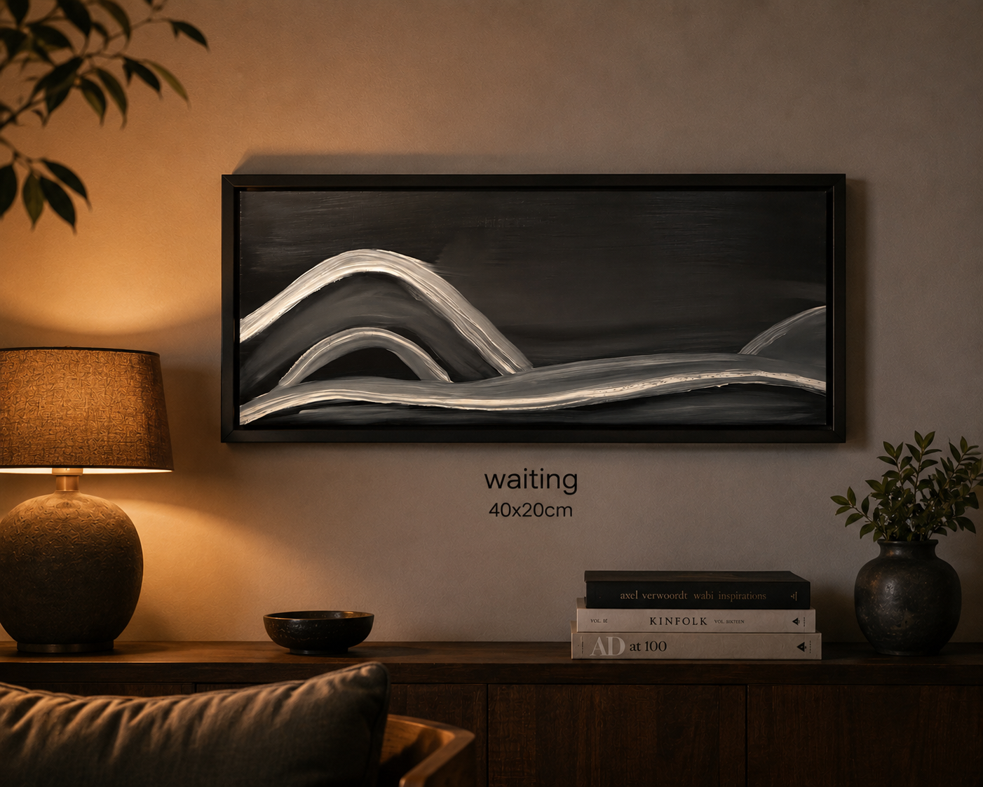 Decorative black and white landscape painting titled "waiting" on a beige wall with a wooden sideboard below, flanked by a textured ceramic table lamp and a dark textured vase with green leafy plant, with a stack of books nearby.