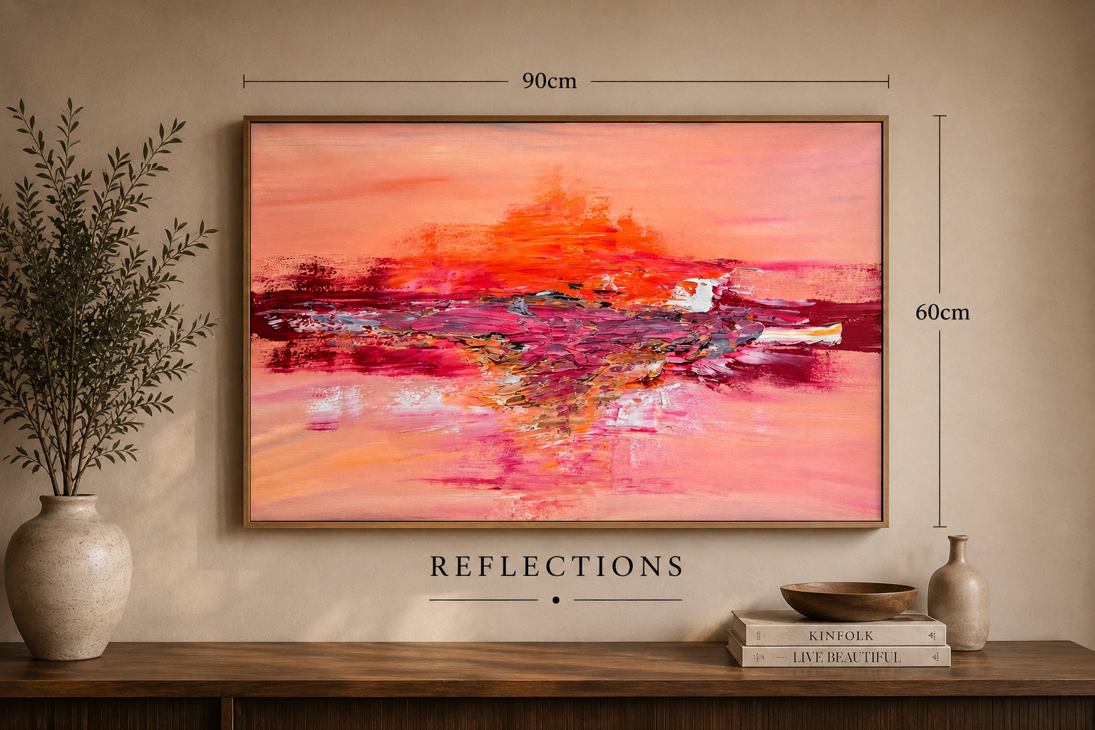Abstract pink, orange, and red watercolor landscape painting hung on a beige wall, with the word 'REFLECTIONS' below, flanked by two thin lines. A potted plant is on the left, and on the right, a small stack of books topped with a wooden bowl and a c