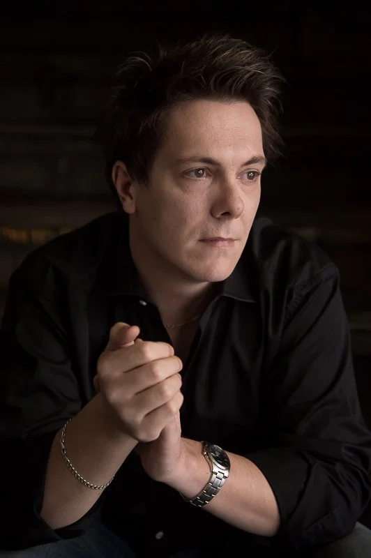 A man with dark hair, wearing a black shirt, is sitting with his hands clasped, looking contemplative, against a dark background.