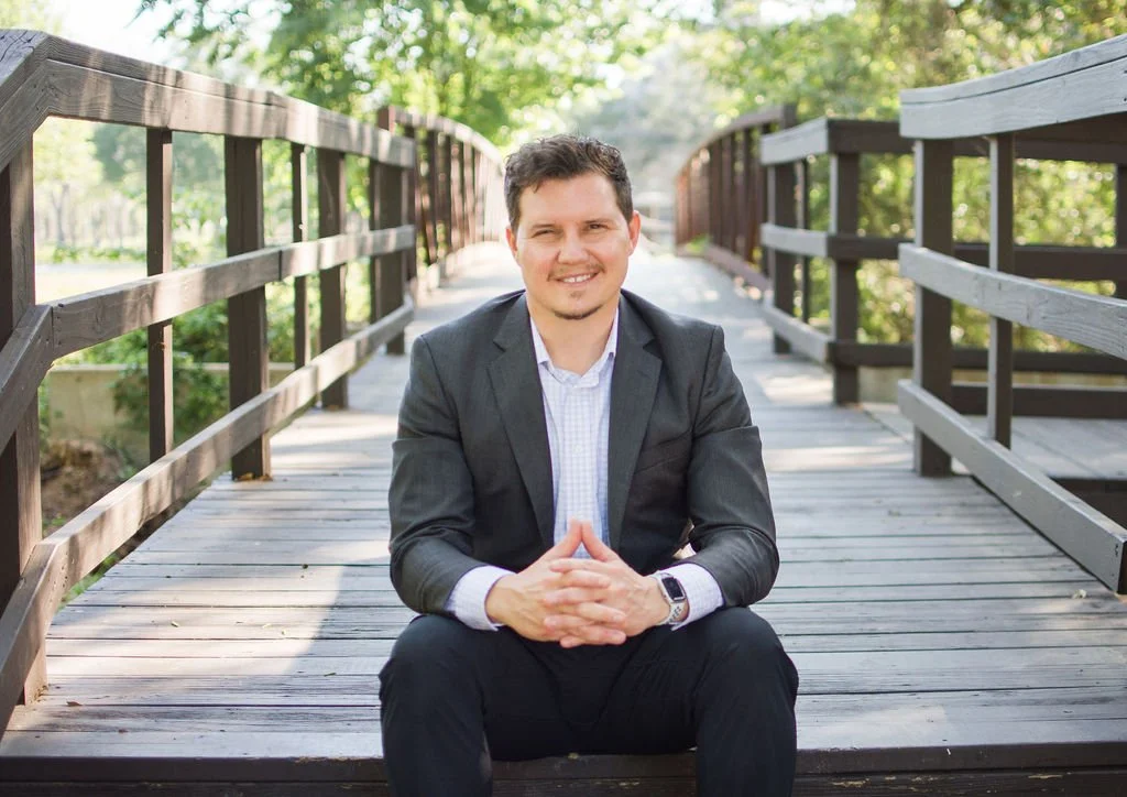 A man in a suit sitting on a wooden bridge outdoors, smiling with hands clasped, surrounded by green trees.