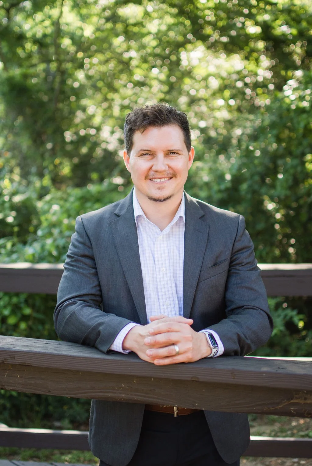 A man in a gray suit and white shirt smiling, with dark hair and facial hair, leaning on a wooden railing outdoors with greenery in the background.
