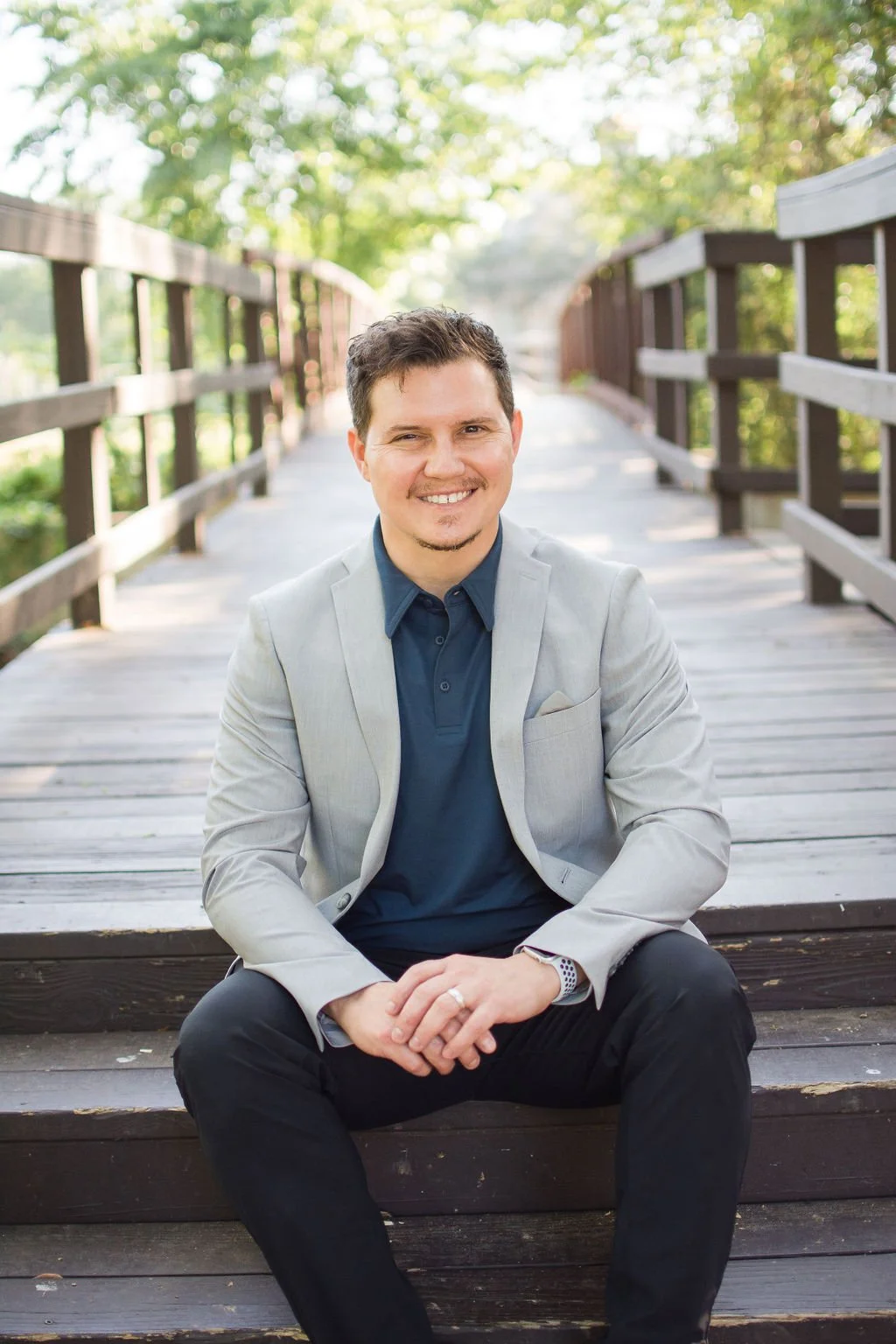 A smiling man in a gray blazer and dark blue polo shirt sitting on a wooden bridge surrounded by greenery.